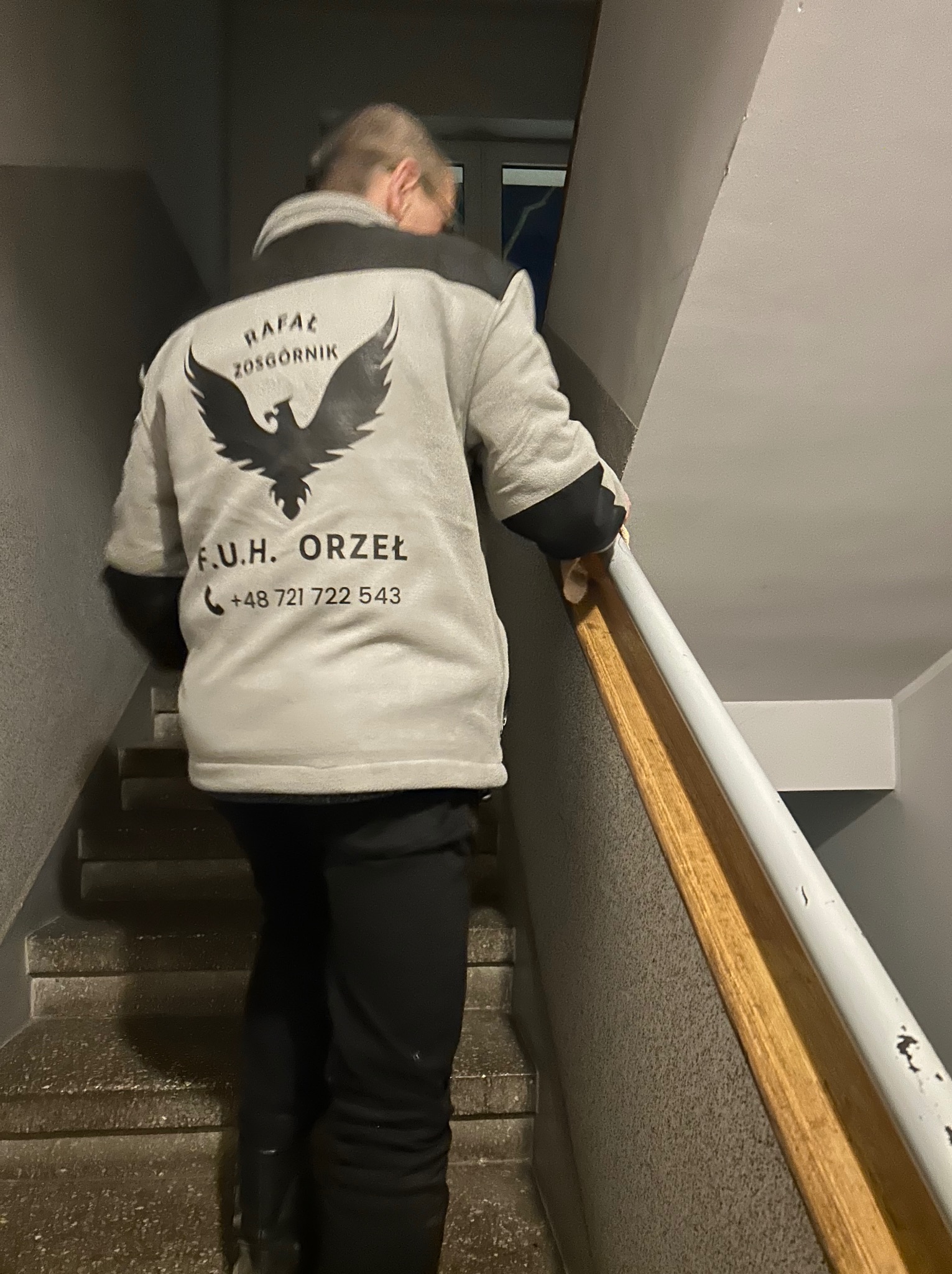 Pracownik firmy sprzątającej w kurtce z logo wchodzi po schodach. Widoczny numer telefonu. Schody betonowe, ściany szare. Ujęcie od tyłu.