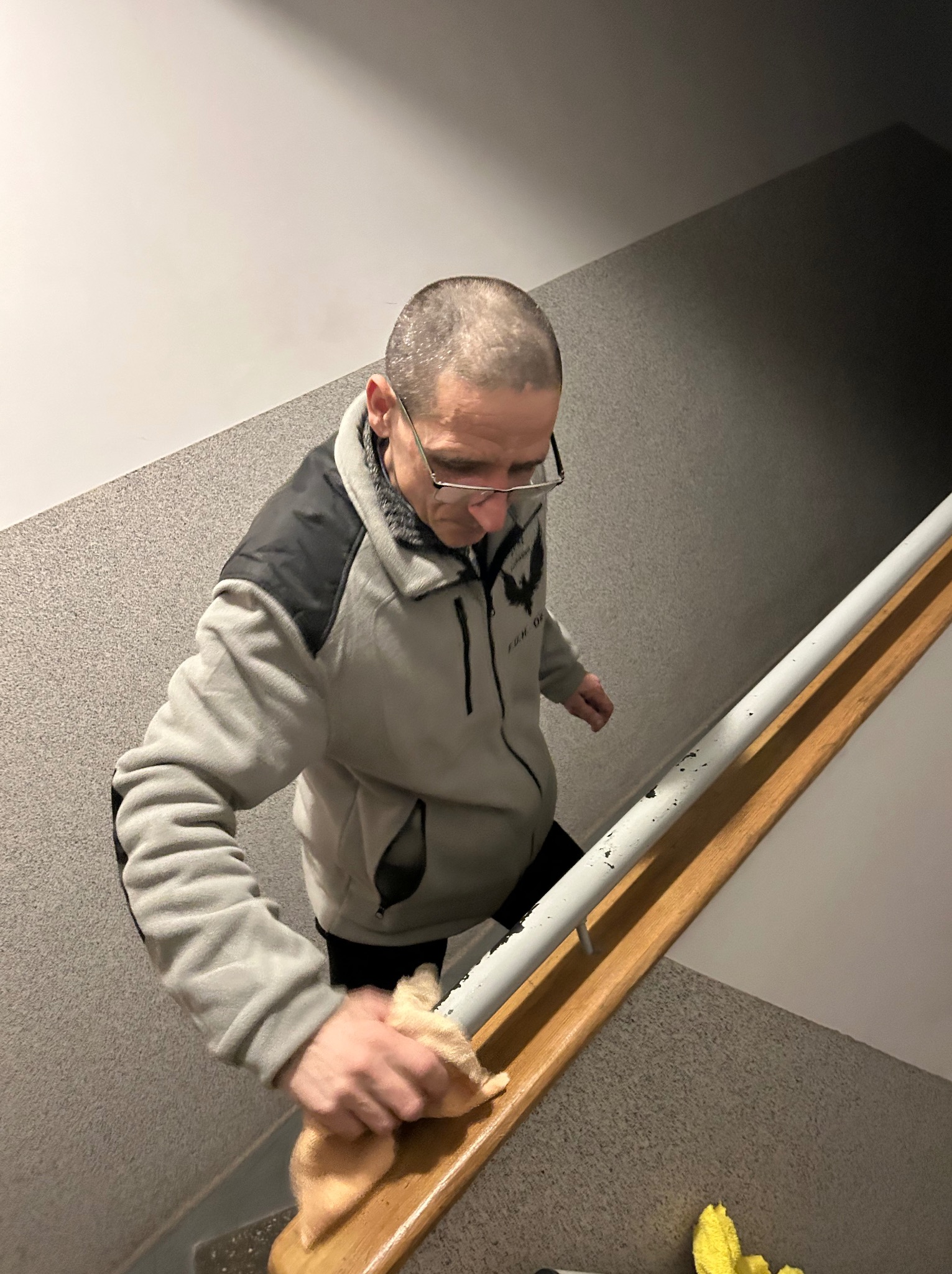 Mężczyzna w okularach czyści drewnianą poręcz schodów szmatką, widok z góry. Na schodach leży żółta ścierka. Szare ściany i schody.
