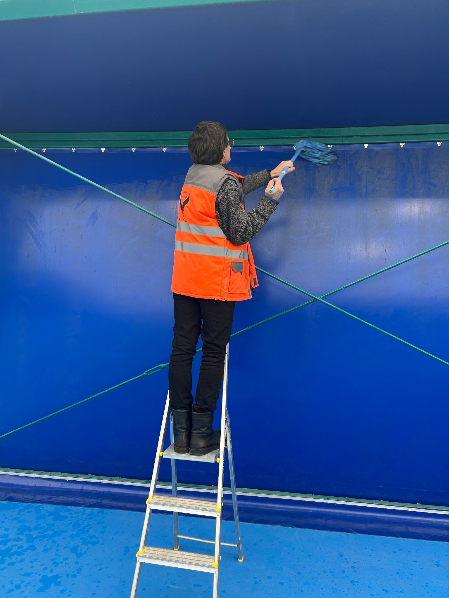 Pracownik w kamizelce na drabinie czyści niebieską, pionową powierzchnię mopem. Widoczne ślady po czyszczeniu. Ujęcie z dołu, podkreślające wysokość czyszczonej powierzchni.
