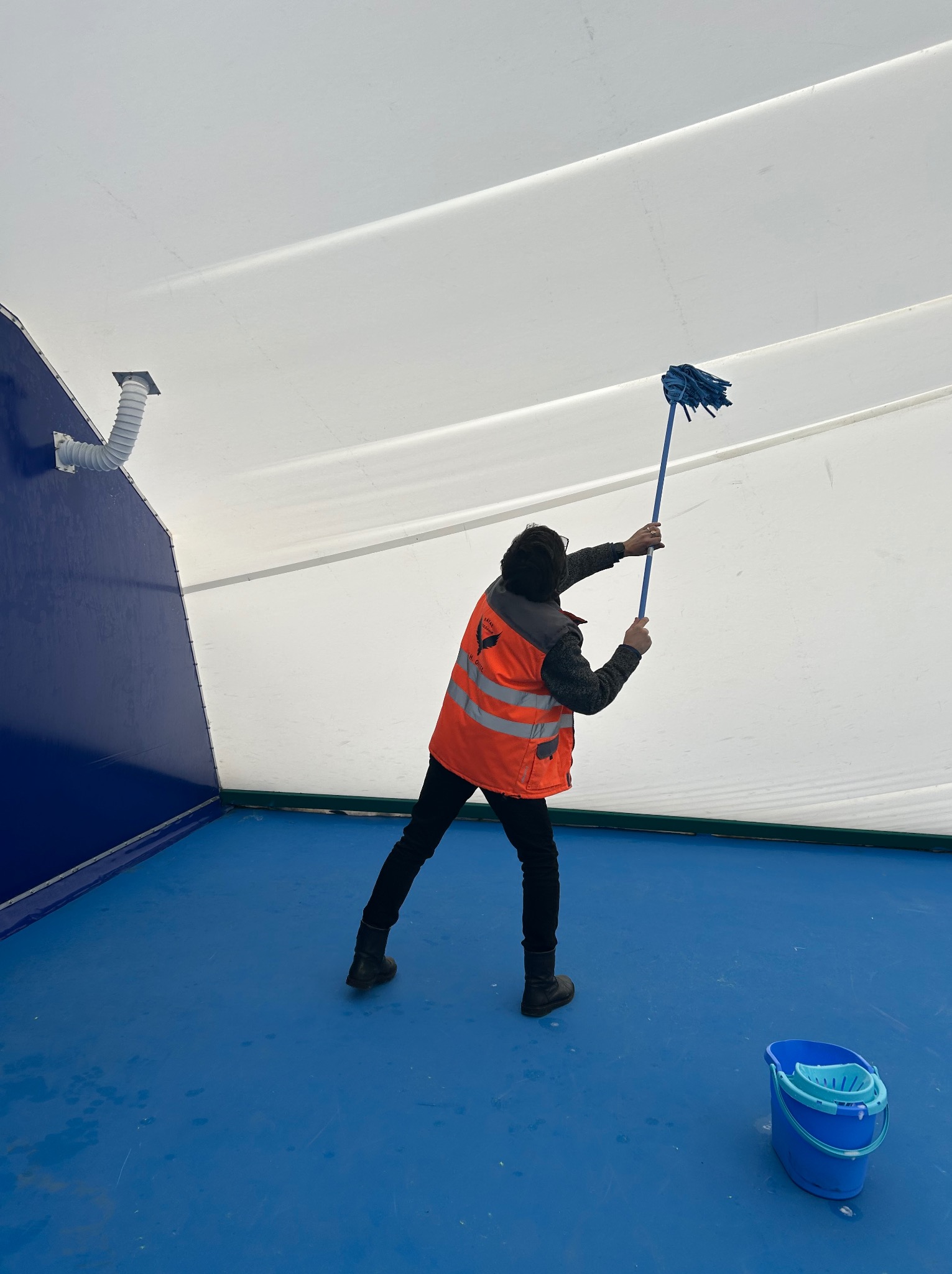 Pracownik w kamizelce odblaskowej myje sufit niebieskim mopem w pomieszczeniu z niebieską podłogą i białymi ścianami. Widoczne wiadro z wodą.