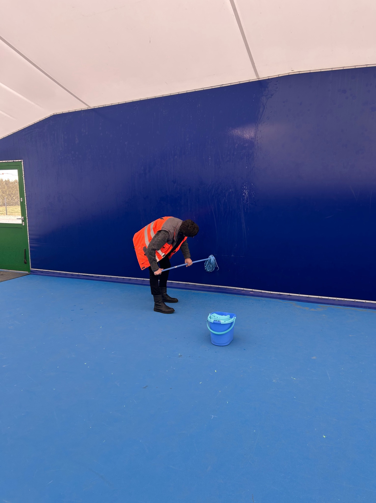 Pracownik w kamizelce odblaskowej myje niebieską ścianę w hali sportowej za pomocą mopa. Obok stoi niebieskie wiadro.
