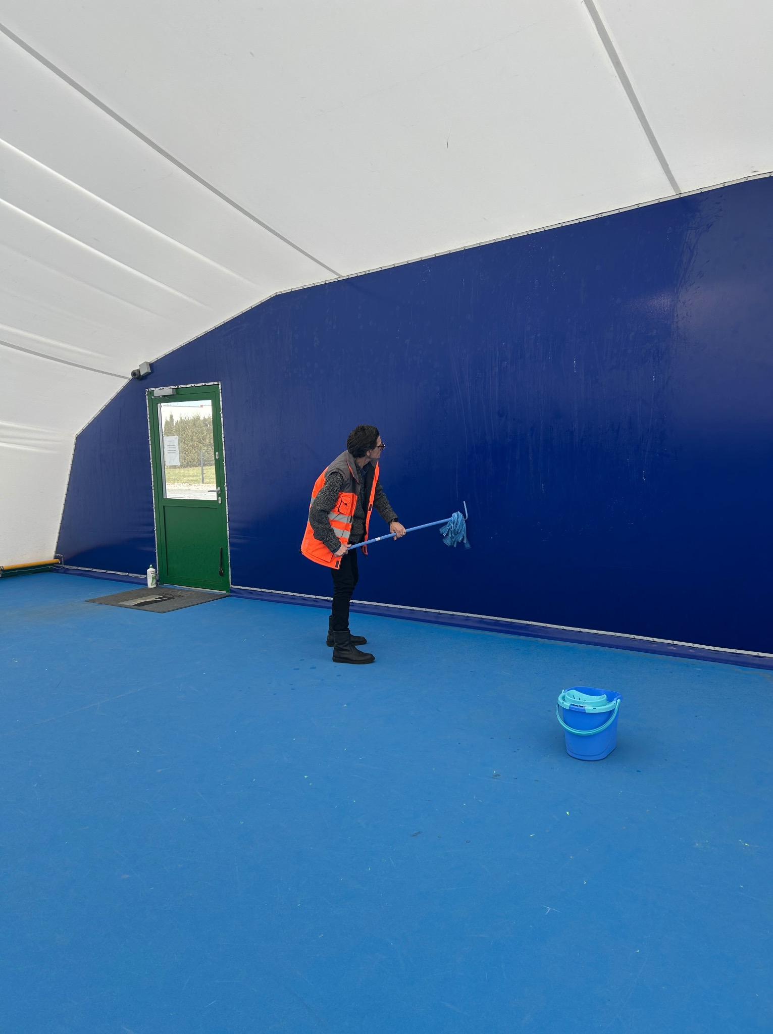 Pracownik w kamizelce odblaskowej myje niebieską ścianę w hali sportowej za pomocą mopa. Obok stoi wiadro. Widoczna brama wejściowa i biały sufit hali.