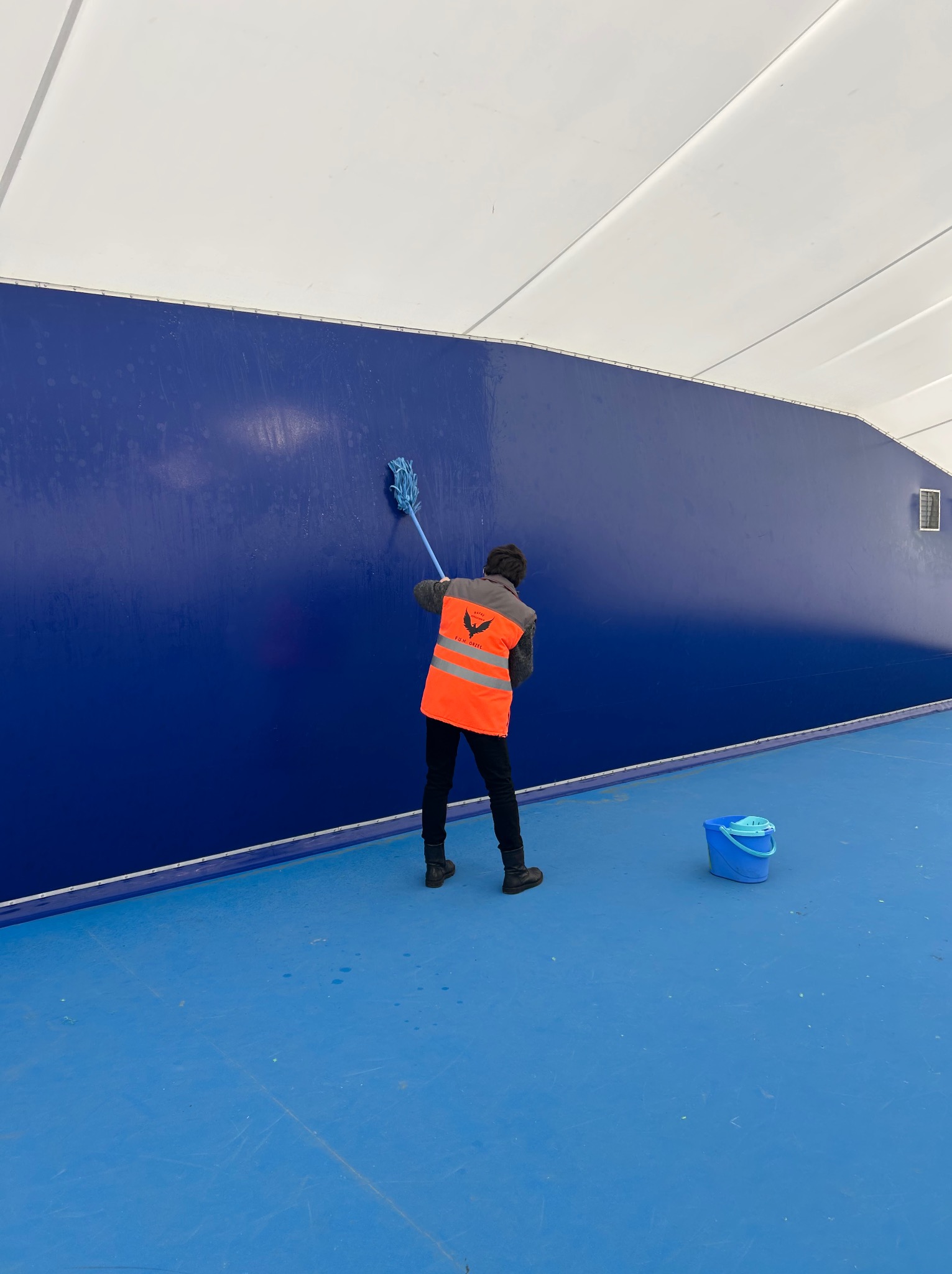 Pracownik w kamizelce myje niebieską ścianę mopem. Widoczne wiadro z wodą. Hala sportowa lub magazyn. Ślady mycia na ścianie.