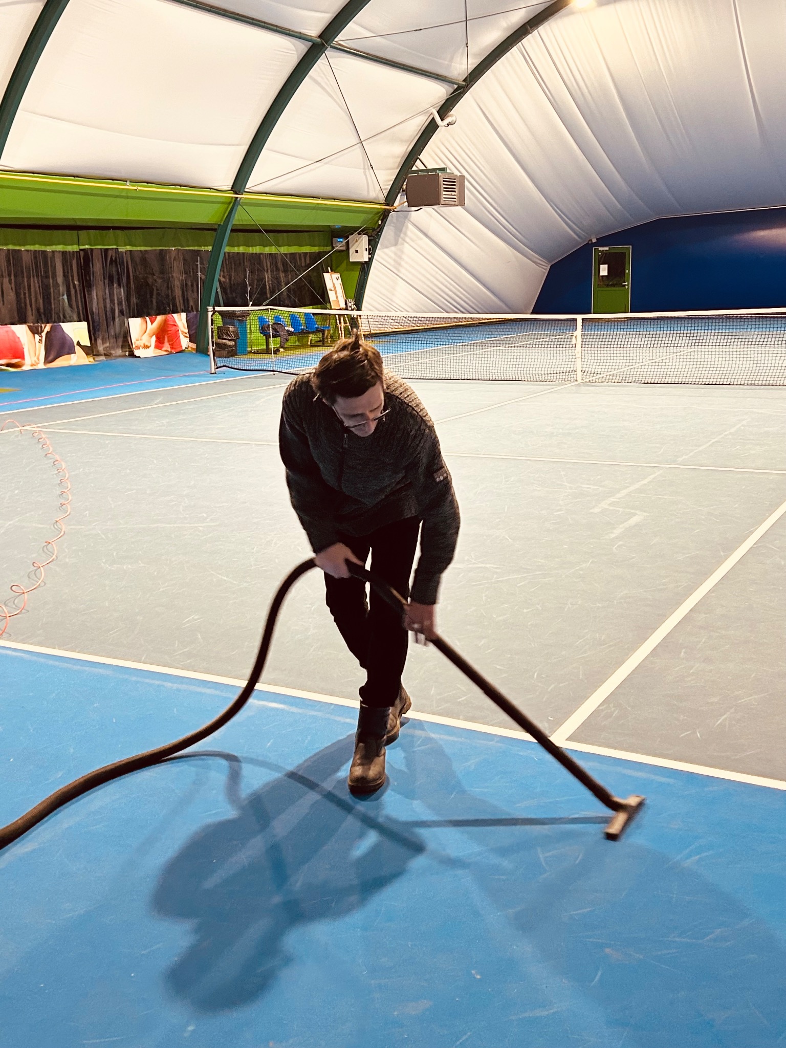 Mężczyzna w okularach odkurza niebieską nawierzchnię kortu tenisowego w hali. Widoczna siatka, oświetlenie i fragmenty wyposażenia sportowego.