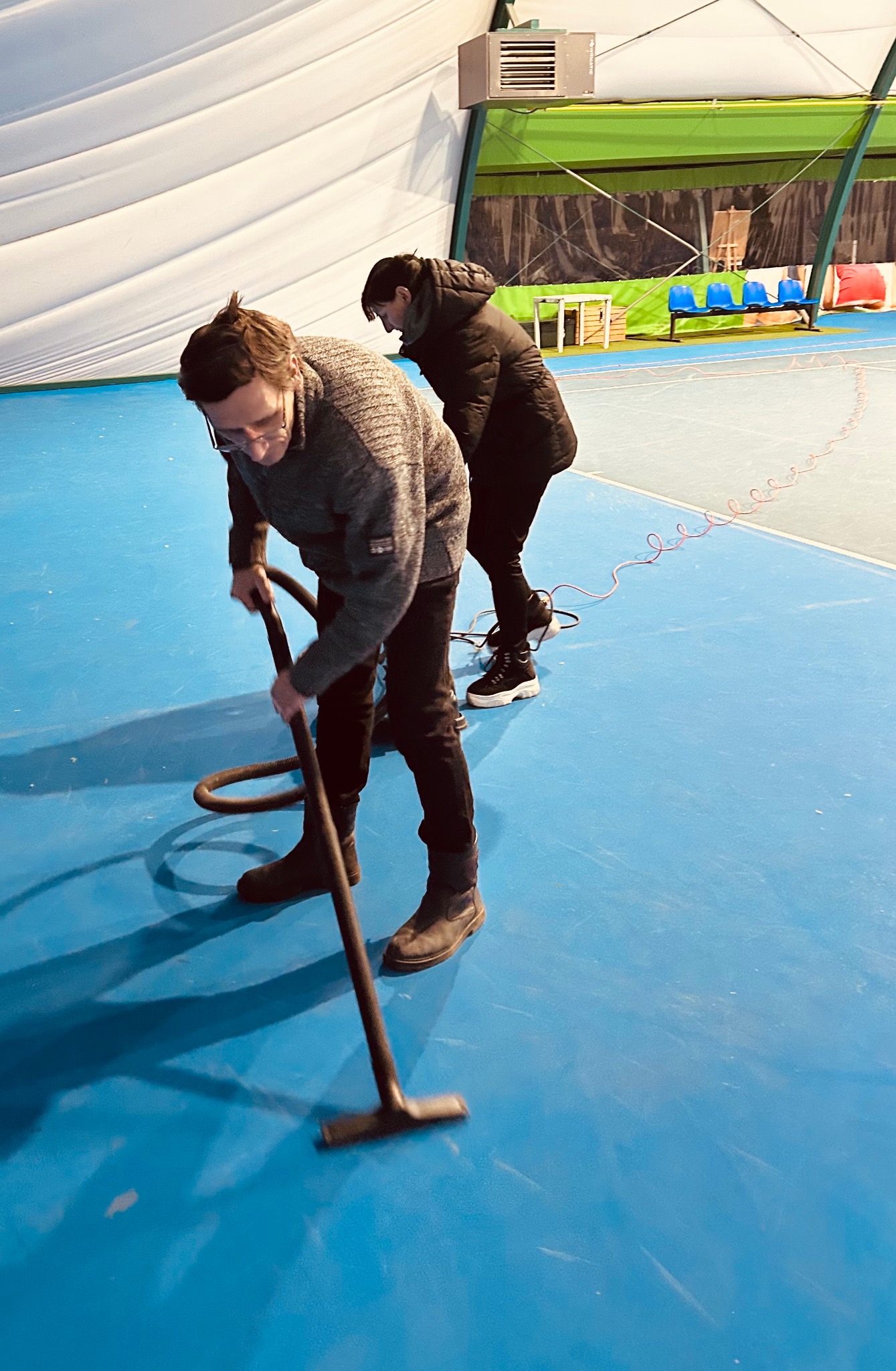 Dwie osoby czyszczą niebieską podłogę hali sportowej za pomocą odkurzacza przemysłowego. W tle widoczne krzesła i konstrukcja hali.