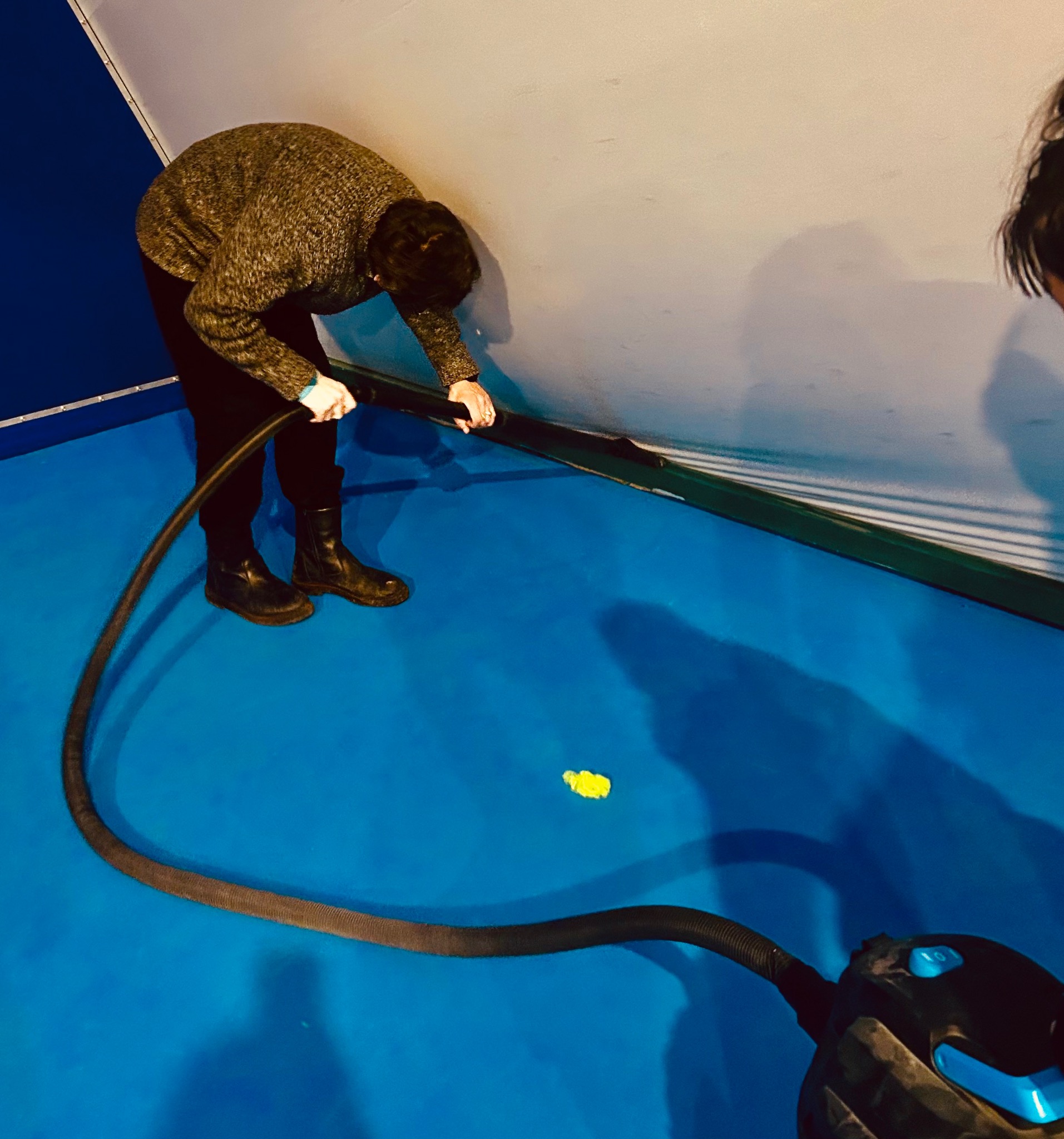 Osoba w brązowym swetrze odkurza niebieską podłogę odkurzaczem z długą rurą, widoczny fragment ściany. Żółty element na podłodze.