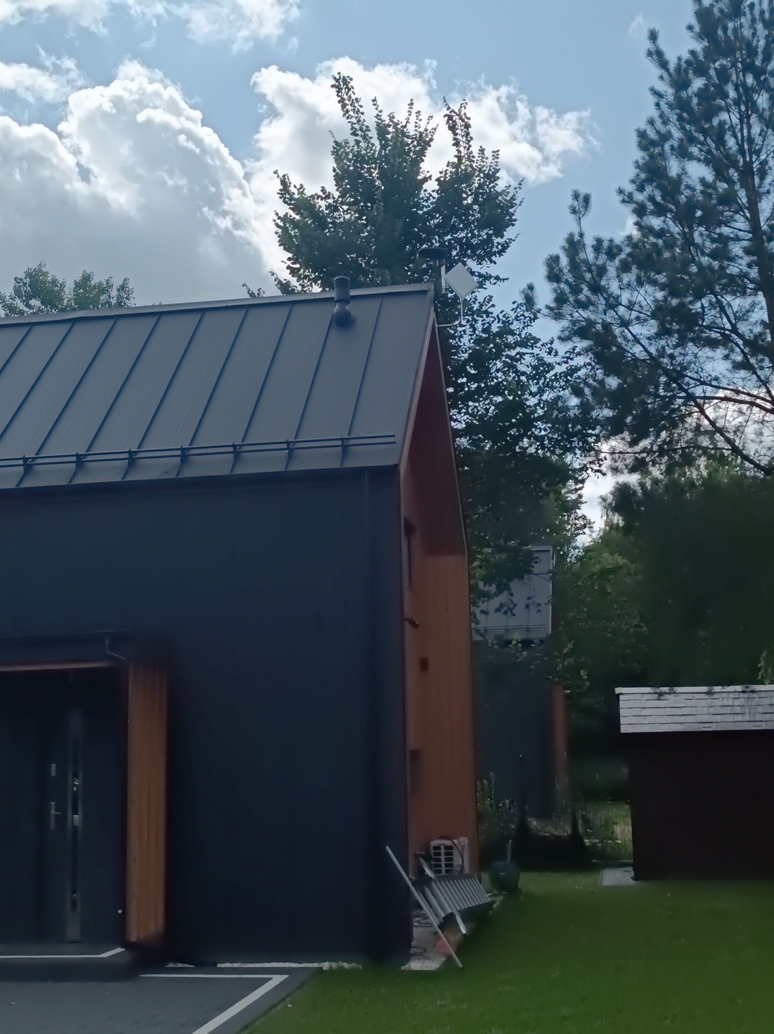 Nowoczesny dom z czarną elewacją i anteną satelitarną na dachu, otoczony zielenią. Widok z dołu, częściowo zasłonięty przez drzewa i krzewy.