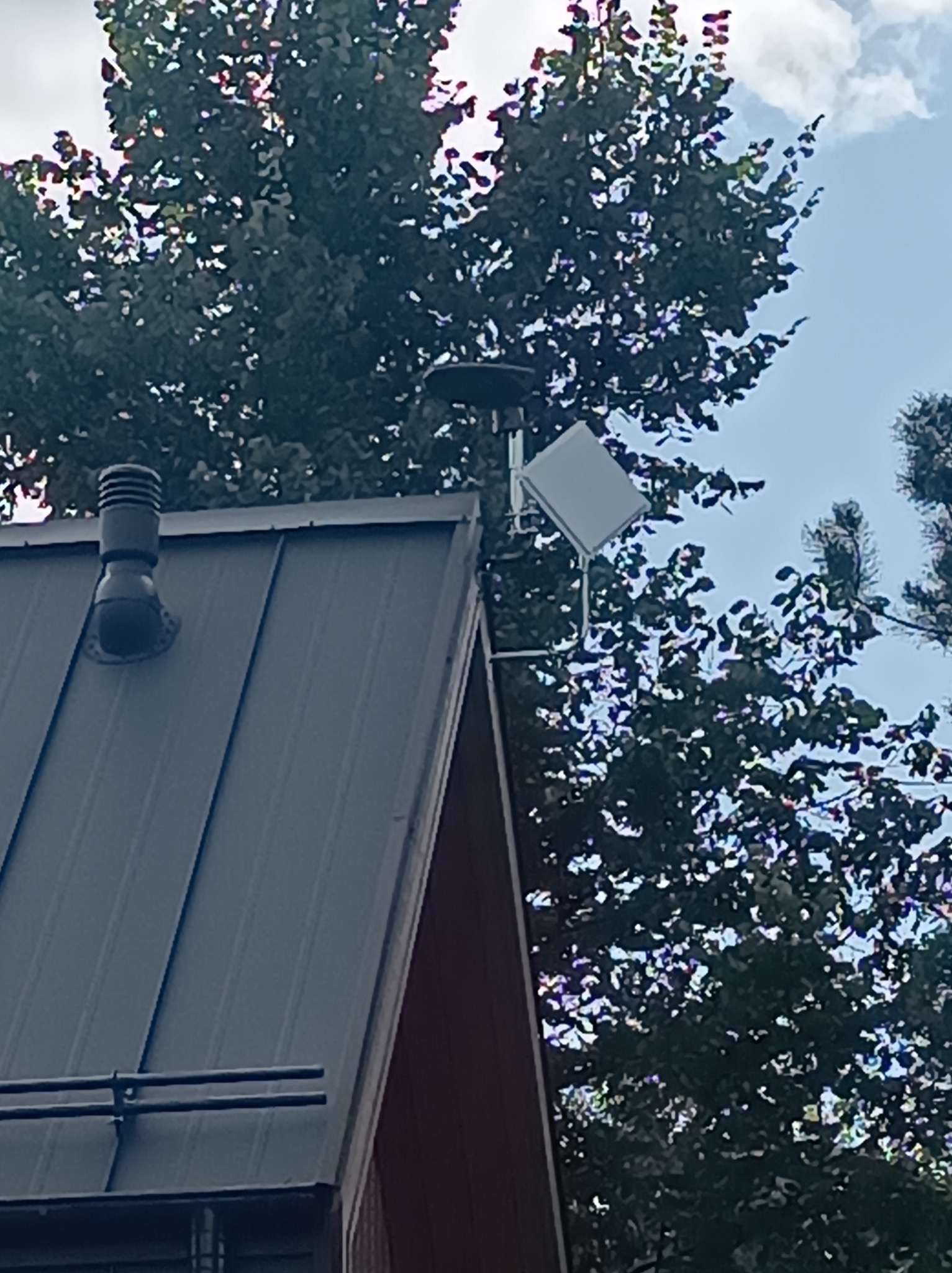 Antena satelitarna zamontowana na metalowym dachu domu jednorodzinnego, widoczna z dołu na tle drzew i fragmentu błękitnego nieba.