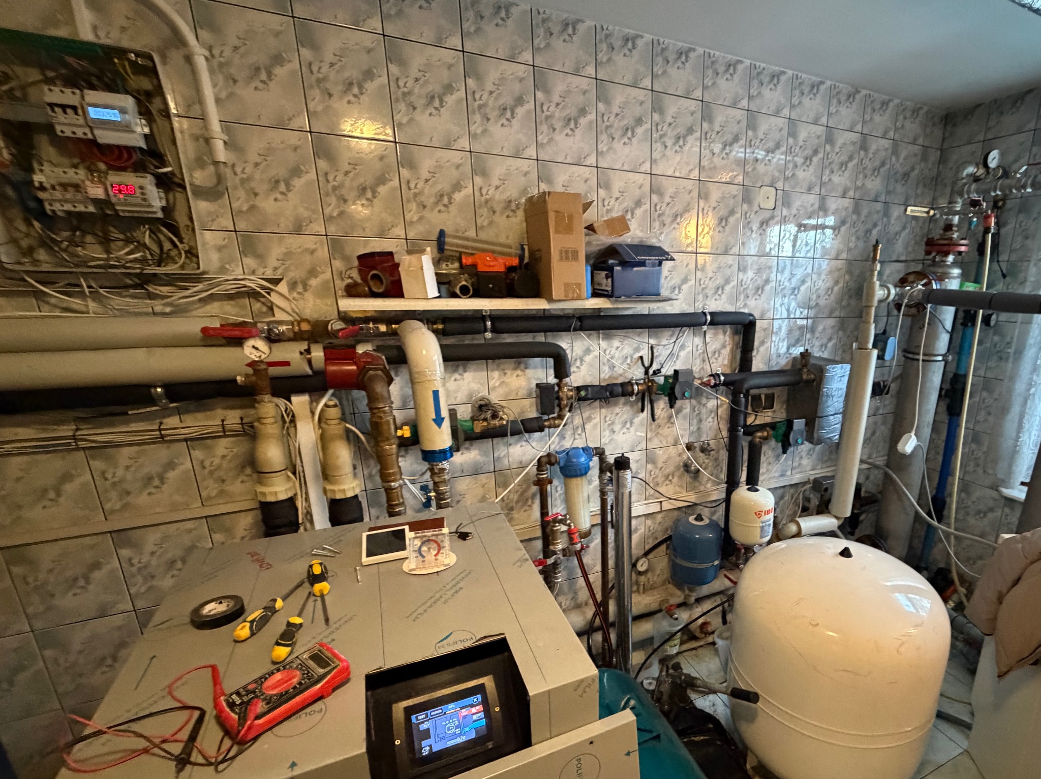 Węzeł cieplny w kotłowni z widocznym sterownikiem, rurami, zaworami i narzędziami. Na ścianie płytki ceramiczne.