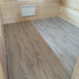 Dawid Markiewicz - Ułożone panele podłogowe w jasnym odcieniu drewna w pomieszczeniu wykończonym boazerią. Widoczny grzejnik na ścianie.