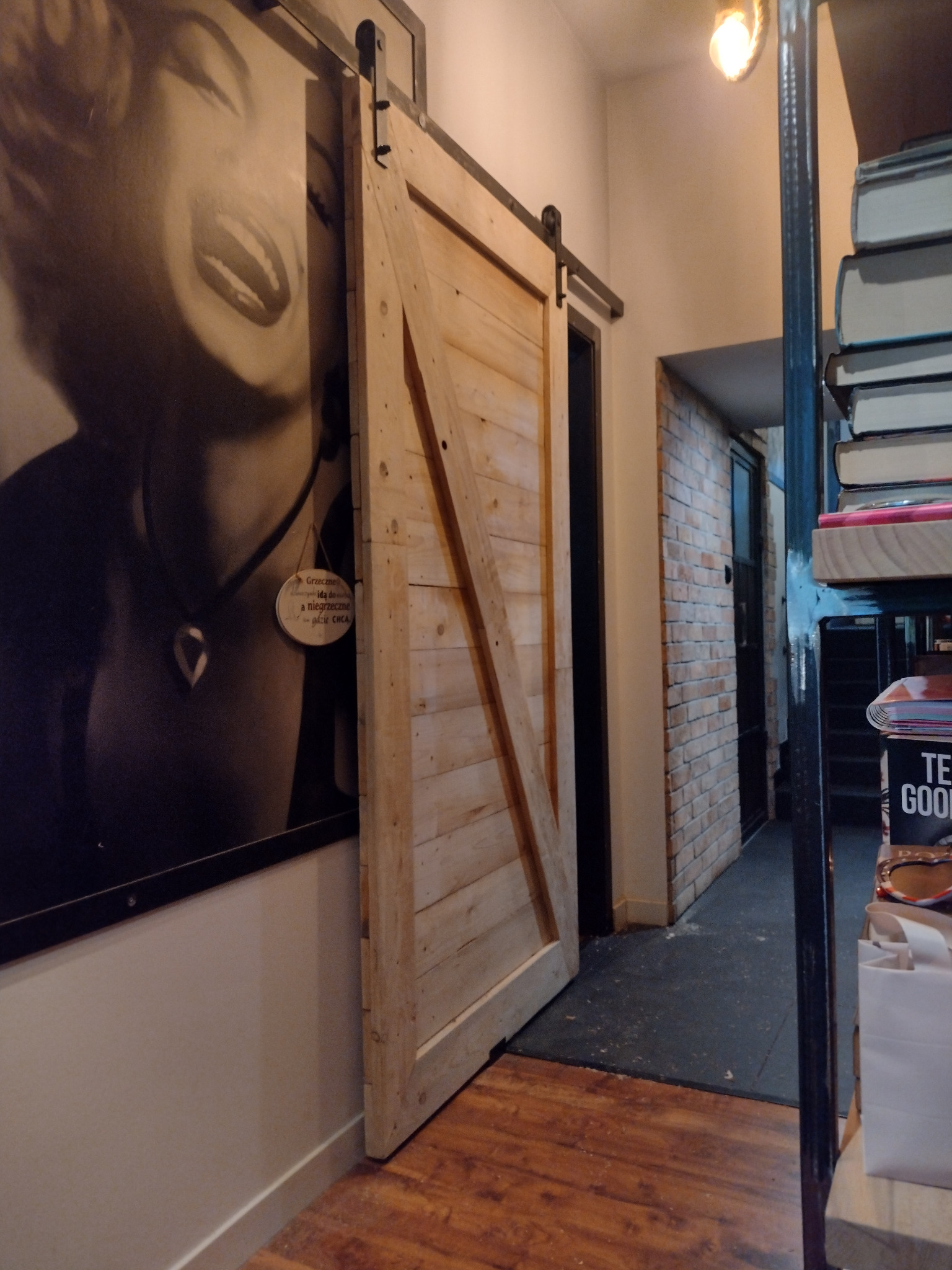 Drewniane drzwi przesuwne w stylu rustykalnym, zamontowane na metalowej szynie, z widoczną grafiką na ścianie przedstawiającą twarz kobiety i fragment ściany z cegły w tle.
