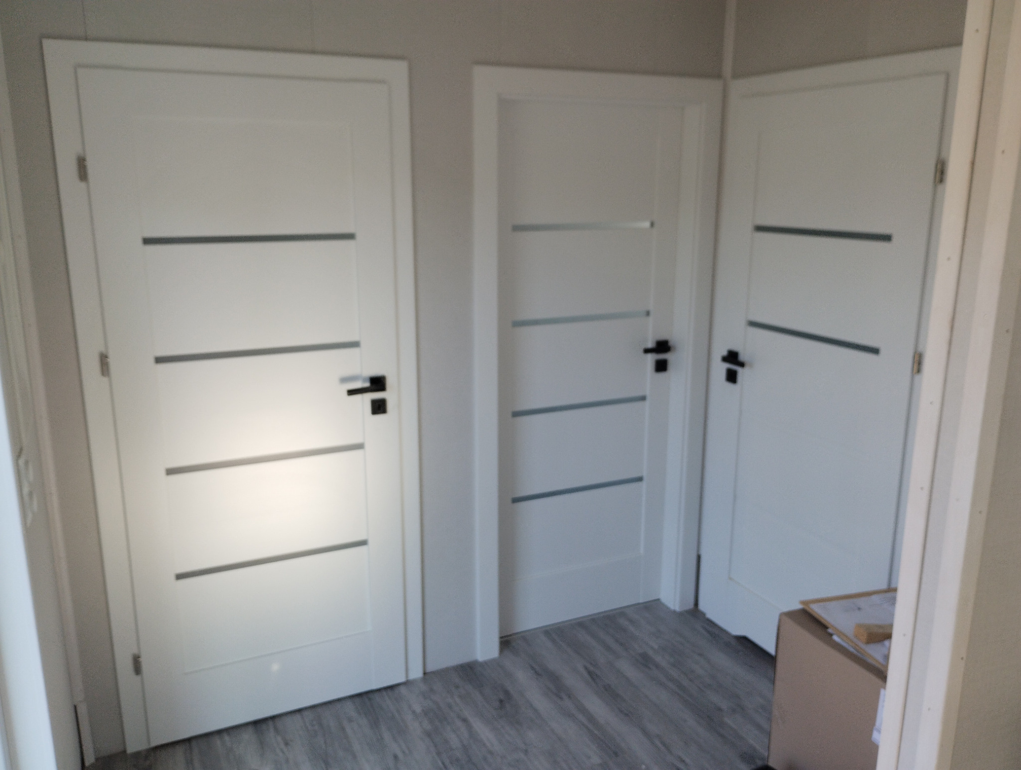 Trzy białe, wewnętrzne drzwi z poziomymi, szarymi paskami i czarnymi klamkami, zamontowane w szarym pomieszczeniu z panelami podłogowymi.