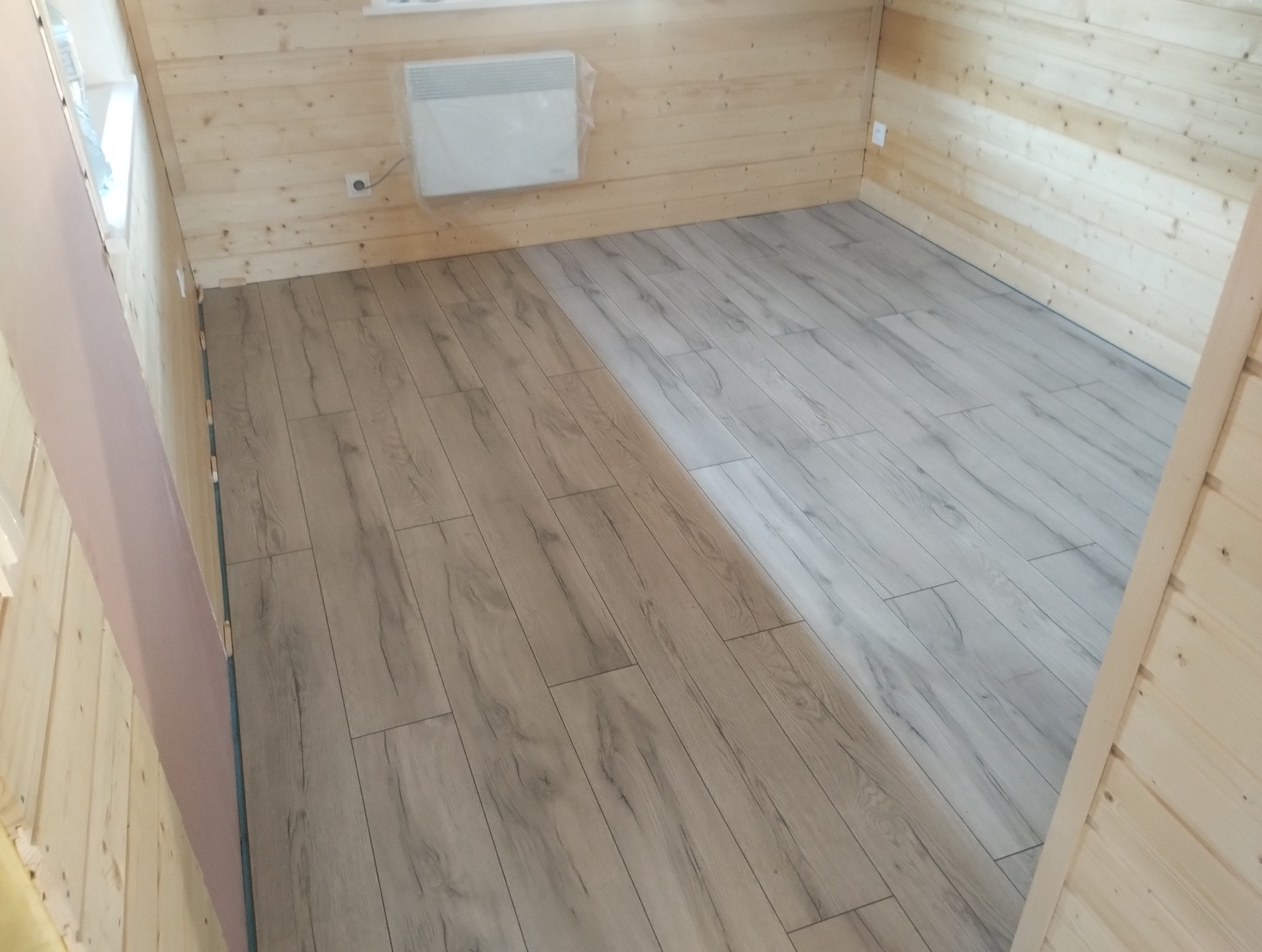 Ułożone panele podłogowe w jasnym odcieniu drewna w pomieszczeniu wykończonym boazerią. Widoczny grzejnik na ścianie.