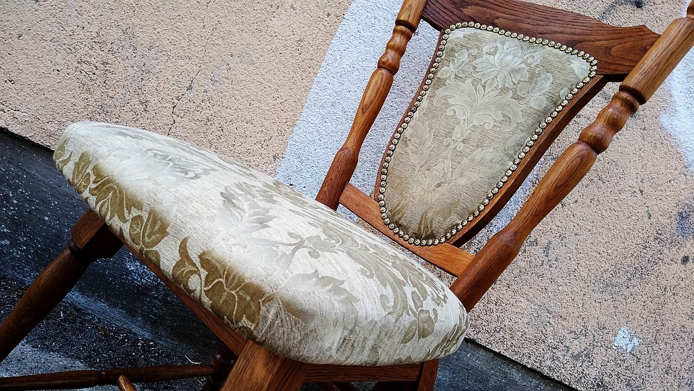 Drewniane krzesło z tapicerowanym siedziskiem i oparciem w kwiatowy wzór, wykończone ozdobnymi ćwiekami, sfotografowane pod kątem na tle ściany.