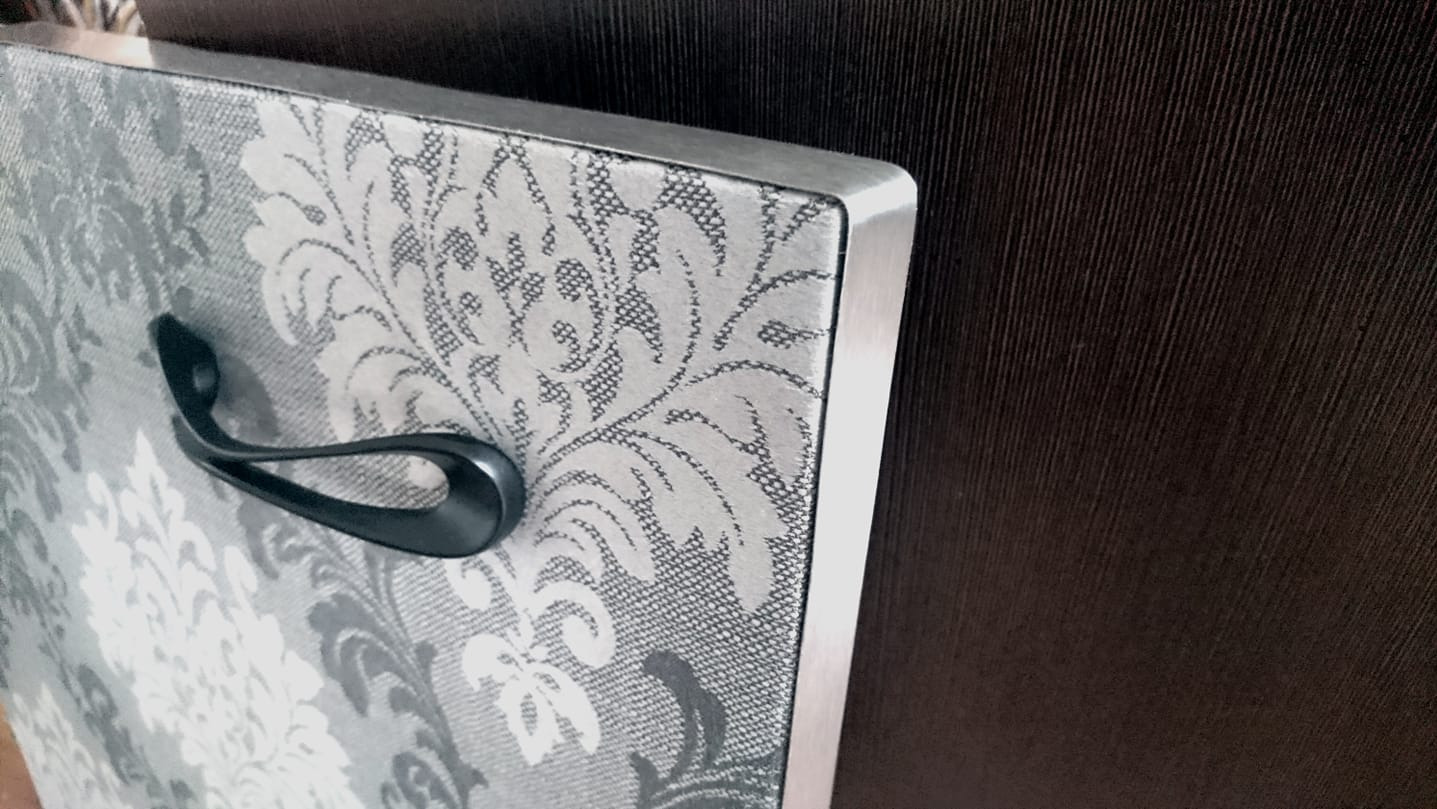 Fragment frontu szafki z ozdobnym, szaro-białym wzorem kwiatowym, metalową ramką i czarnym, nowoczesnym uchwytem, na tle ciemnej, drewnopodobnej płyty.