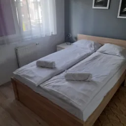 Pokój hotelowy z dwoma łóżkami pojedynczymi, białymi pościelami, zwiniętymi ręcznikami, grzejnikiem pod oknem i minimalistycznymi obrazami na szarej ścianie.