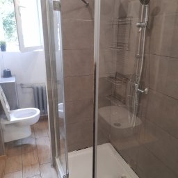 Clean Vibe Bartłomiej Wierzbicki - Widok na kabinę prysznicową z szarymi płytkami, metalowymi półkami i zestawem prysznicowym, widoczny fragment toalety i kaloryfera w tle, ślady wilgoci na podłodze sugerują niedawne użycie.