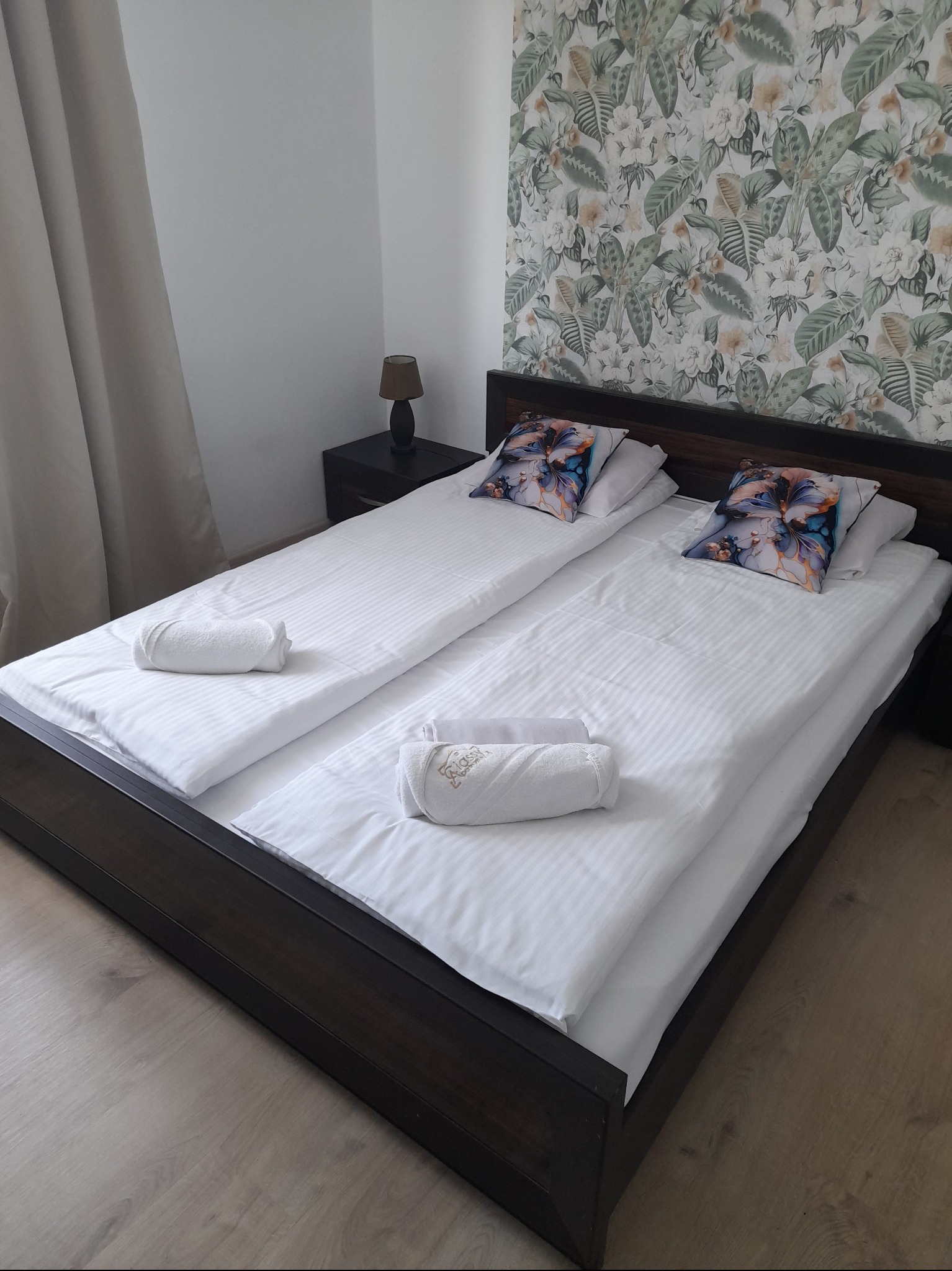 Pokój hotelowy z dwoma łóżkami z ciemnego drewna, białą pościelą w paski, wzorzystymi poduszkami i ręcznikami z logo, na ścianie tapeta w tropikalne liście.