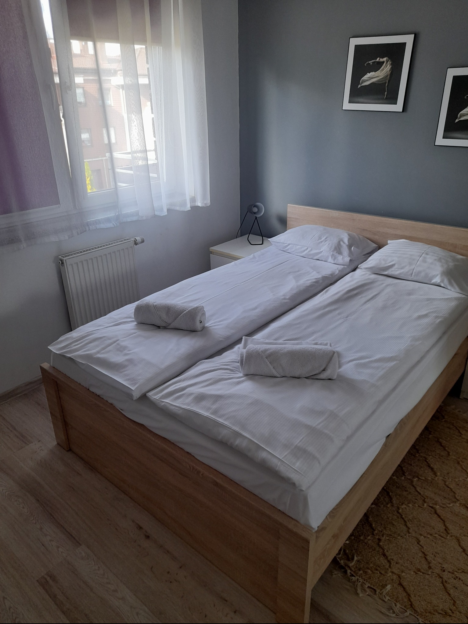 Pokój hotelowy z dwoma łóżkami pojedynczymi, białymi pościelami, zwiniętymi ręcznikami, grzejnikiem pod oknem i minimalistycznymi obrazami na szarej ścianie.