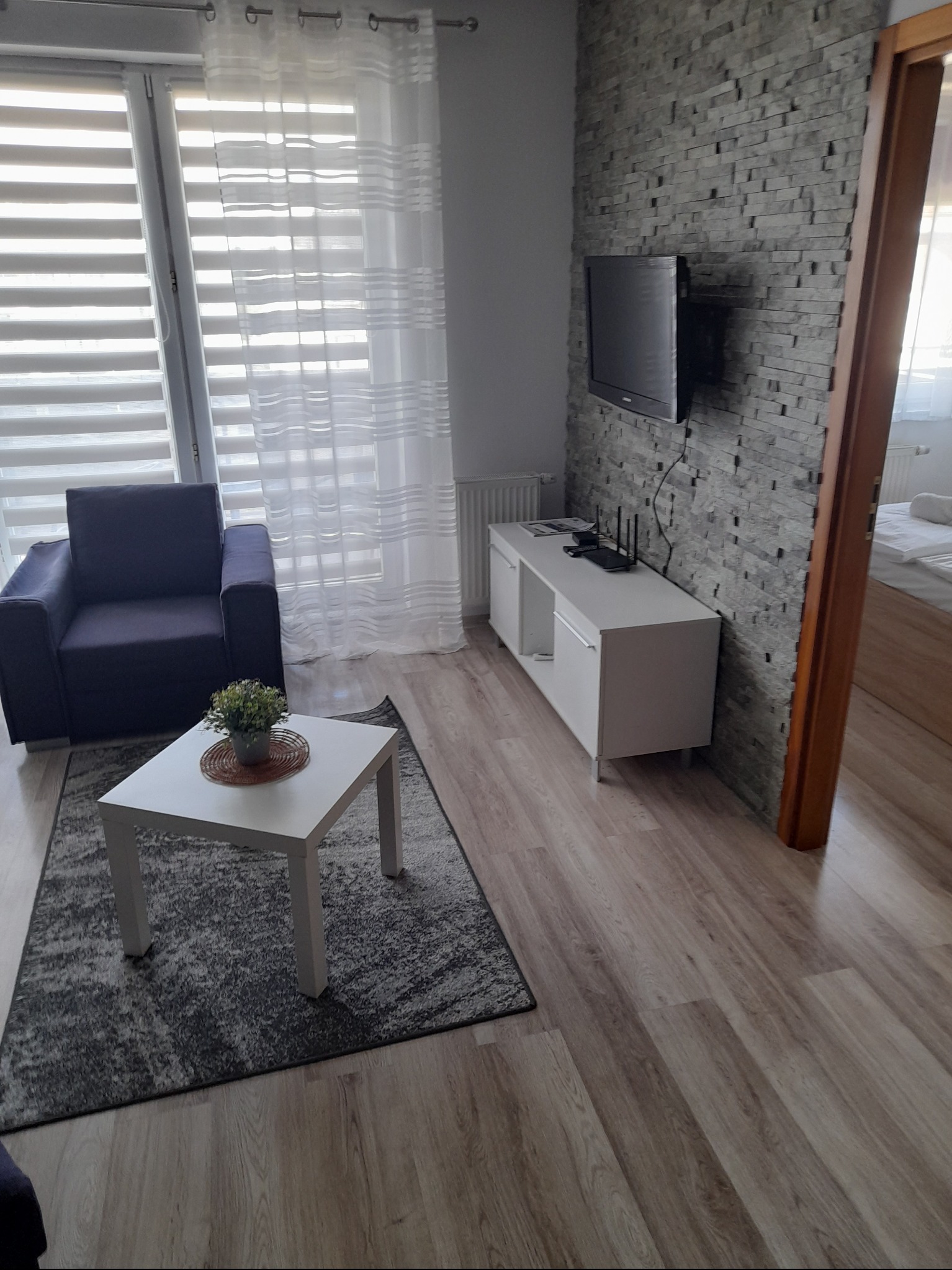 Jasny pokój dzienny z szarym dywanem, ciemnofioletowym fotelem i białą szafką RTV na tle ściany z szarej cegły dekoracyjnej. Na stoliku kawowym roślina doniczkowa.