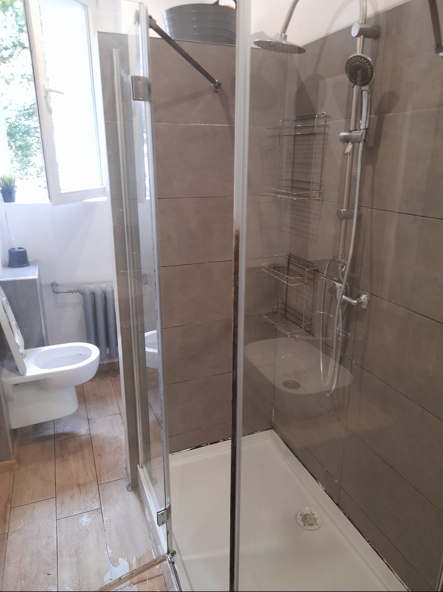 Widok na kabinę prysznicową z szarymi płytkami, metalowymi półkami i zestawem prysznicowym, widoczny fragment toalety i kaloryfera w tle, ślady wilgoci na podłodze sugerują niedawne użycie.