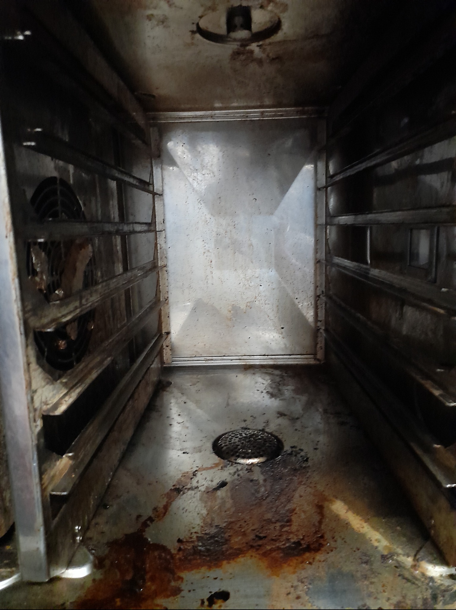 Wnętrze brudnego piekarnika konwekcyjnego z widocznymi zaschniętymi resztkami jedzenia i tłuszczu na ścianach, dnie oraz ruszcie wentylatora.