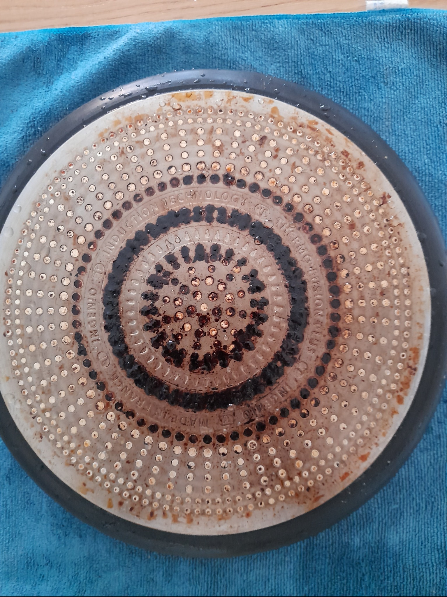 Spód brudnej patelni z technologią indukcyjną, widoczne przypalone resztki jedzenia i napis 'Induction Technology' na niebieskiej ścierce.