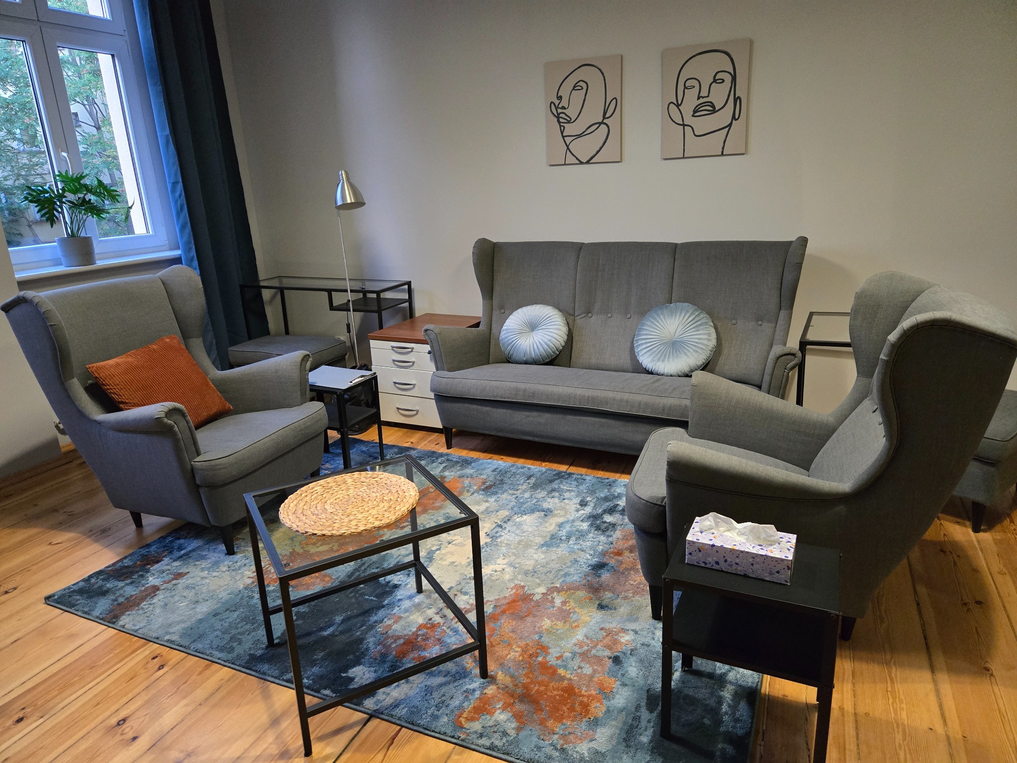 Pokój z szarymi fotelami uszakami, sofą i stolikami na tle ściany z abstrakcyjnymi obrazami linii twarzy, dywan w odcieniach niebieskiego i brązu na drewnianej podłodze.