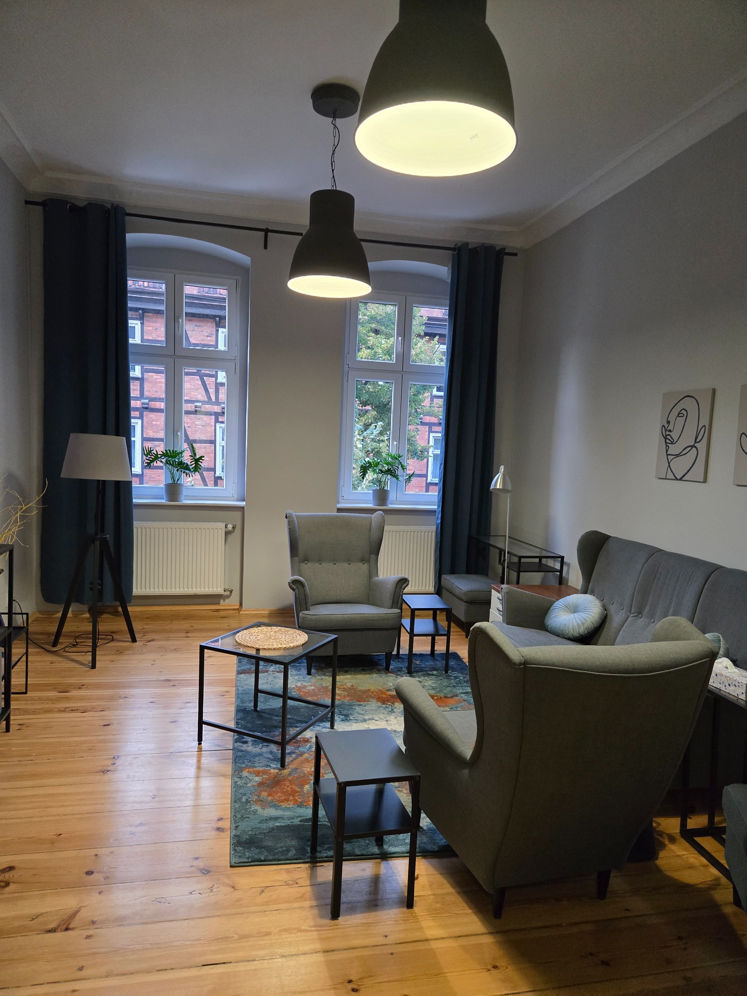 Jasne, przestronne wnętrze pokoju z dwoma oknami z widokiem na ceglany budynek, szare fotele typu uszak, kanapa, stoliki kawowe i lampy wiszące.