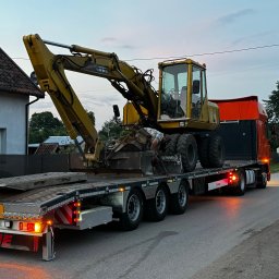 Elektro-Art Marcin Pycek - Żółta koparka na niskopodwoziowej naczepie ciężarówki, pracownik w koszuli w hawajskim stylu przy łyżce koparki, transport maszyny budowlanej.