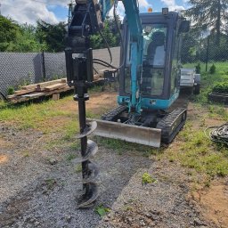 Elektro-Art Marcin Pycek - Niebieska minikoparka z wiertnicą hydrauliczną na nierównym terenie z gruzem i trawą, przygotowana do wiercenia otworów pod instalacje.