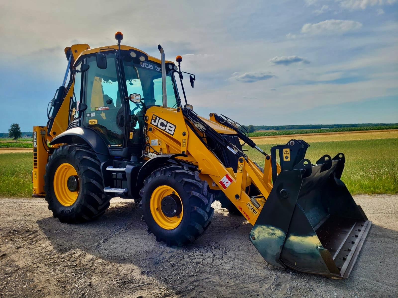 Żółto-czarna koparko-ładowarka JCB 3CX zaparkowana na żwirowej drodze, z widocznym polem w tle pod błękitnym niebem z lekkimi chmurami.