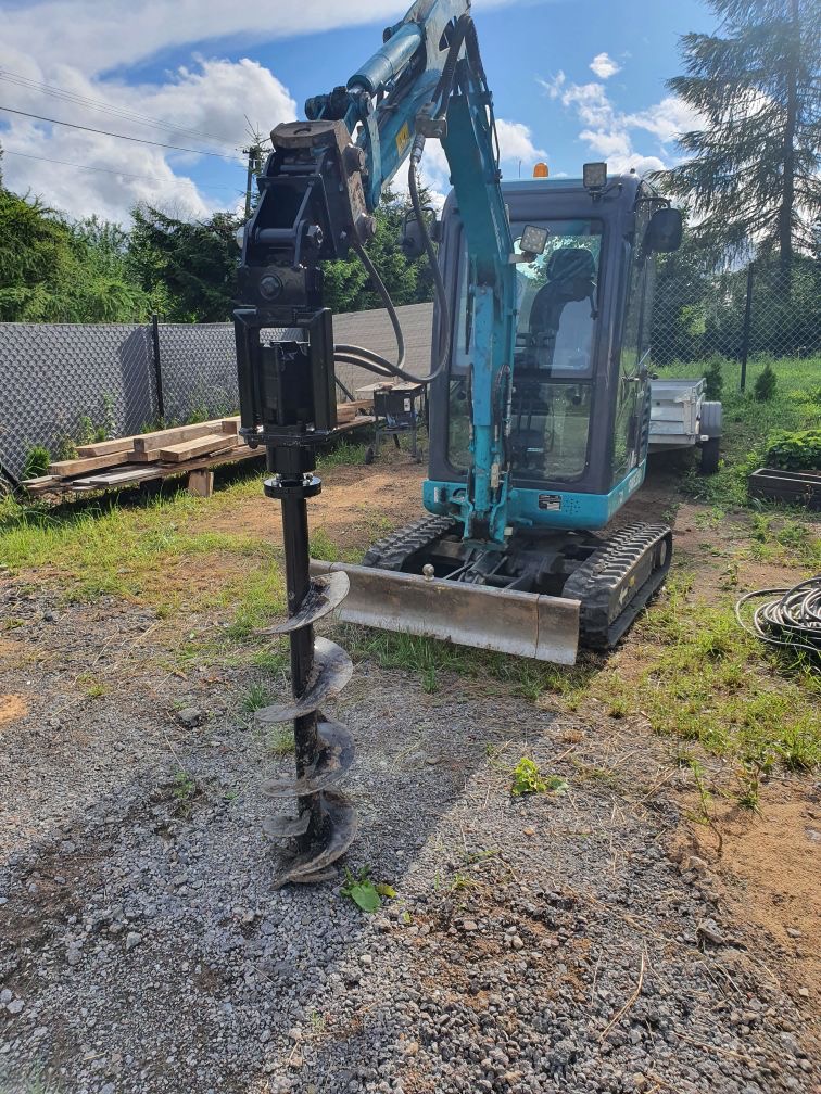 Niebieska minikoparka z wiertnicą hydrauliczną na nierównym terenie z gruzem i trawą, przygotowana do wiercenia otworów pod instalacje.