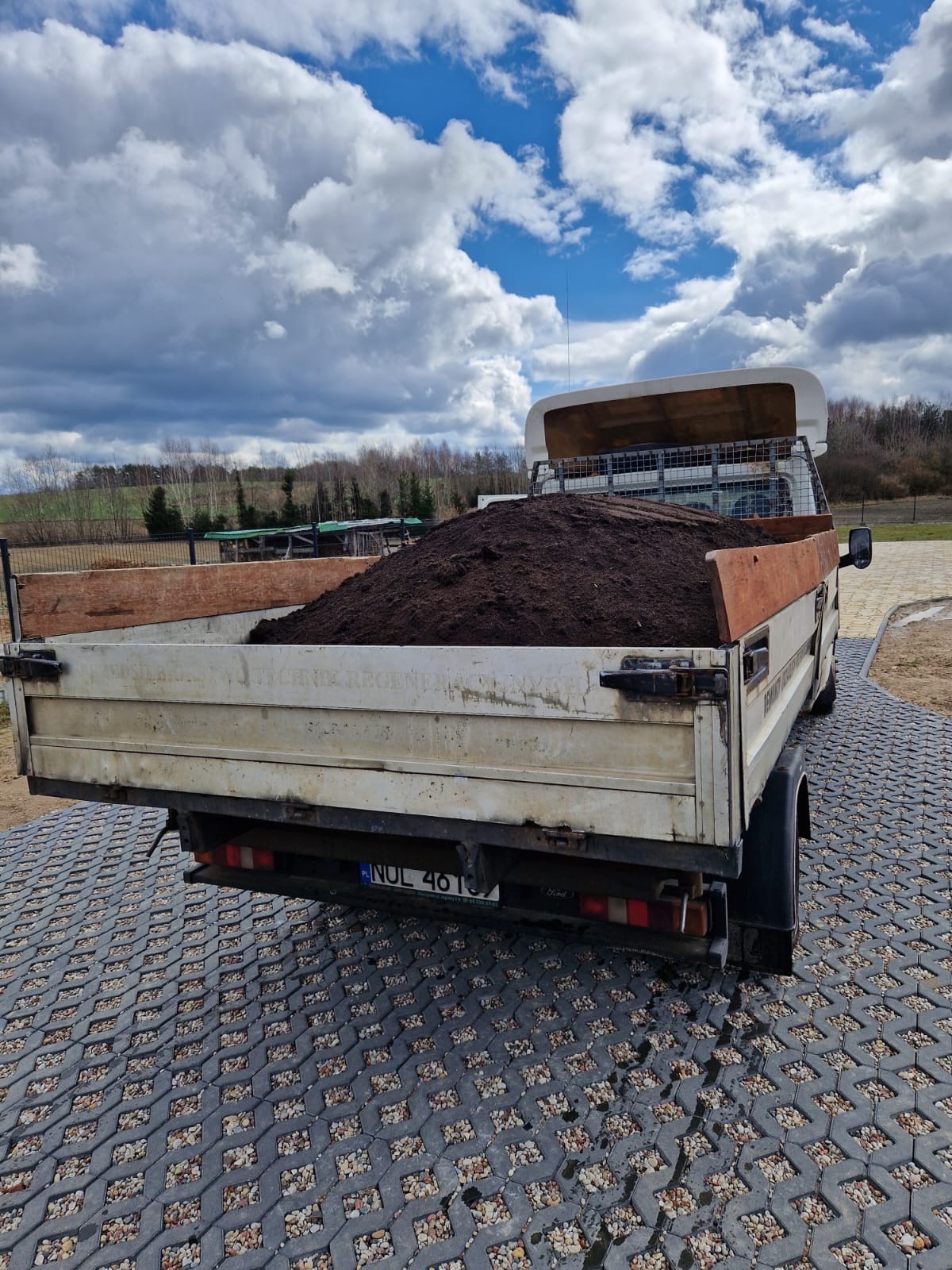 Tył białego samochodu dostawczego z przyczepą wypełnioną ziemią, zaparkowanego na ażurowej kostce brukowej w Olsztynku, na tle pochmurnego nieba.