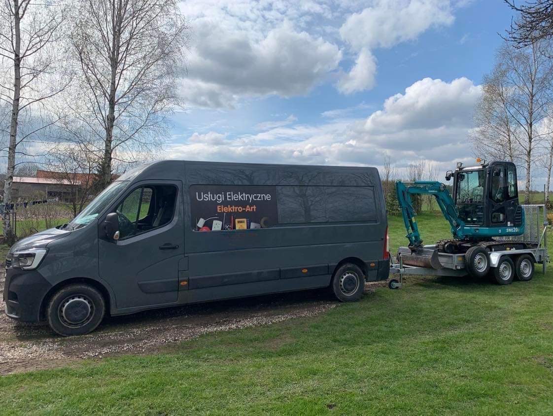 Szary van z reklamą usług elektrycznych 'Elektro-Art' ciągnie na przyczepie mini-koparkę na tle zielonej trawy i błękitnego nieba z chmurami.