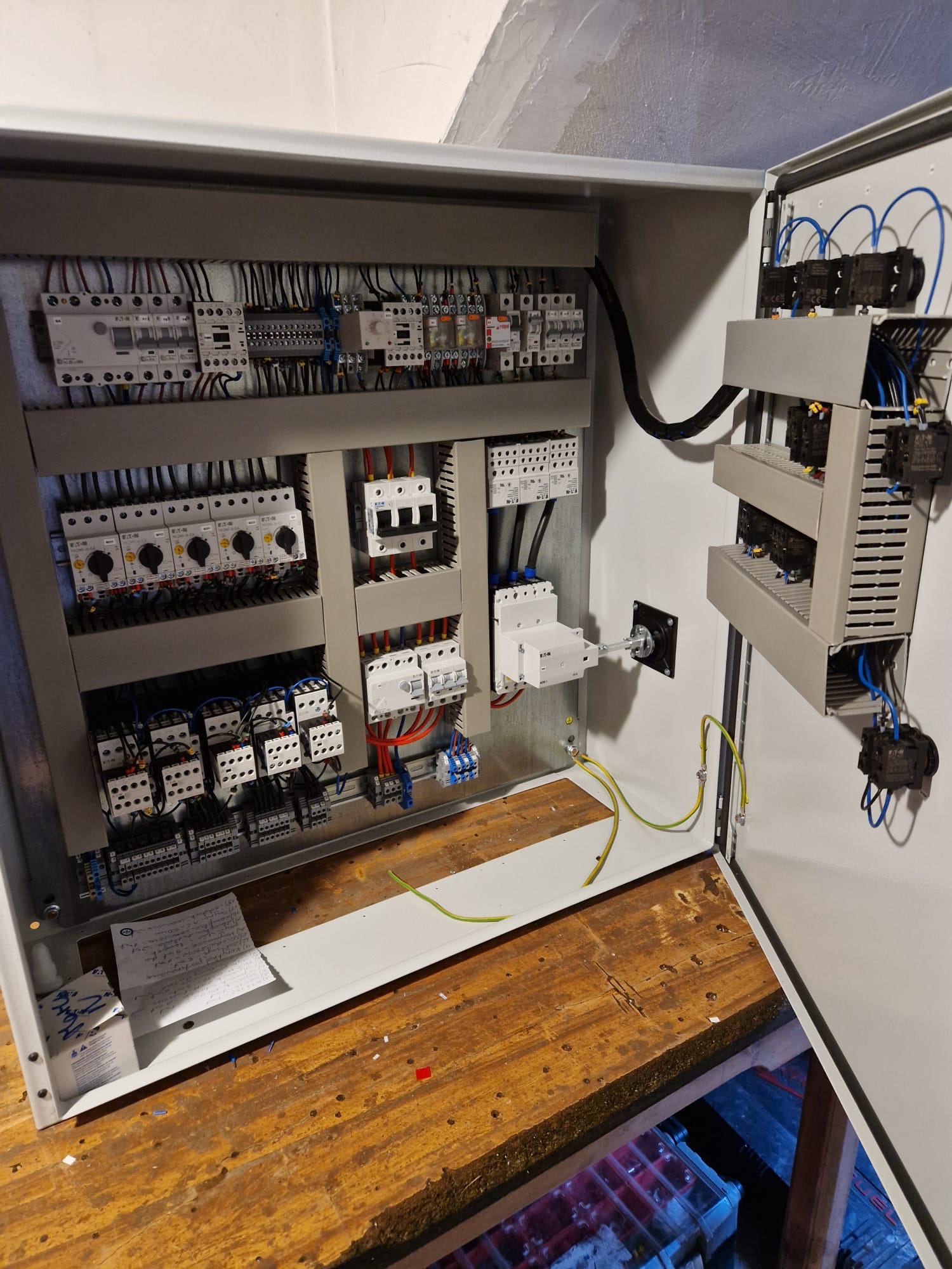 Otwarta szafa sterownicza z widocznym okablowaniem, bezpiecznikami, stycznikami i wyłącznikami, zamontowana na drewnianej powierzchni roboczej.
