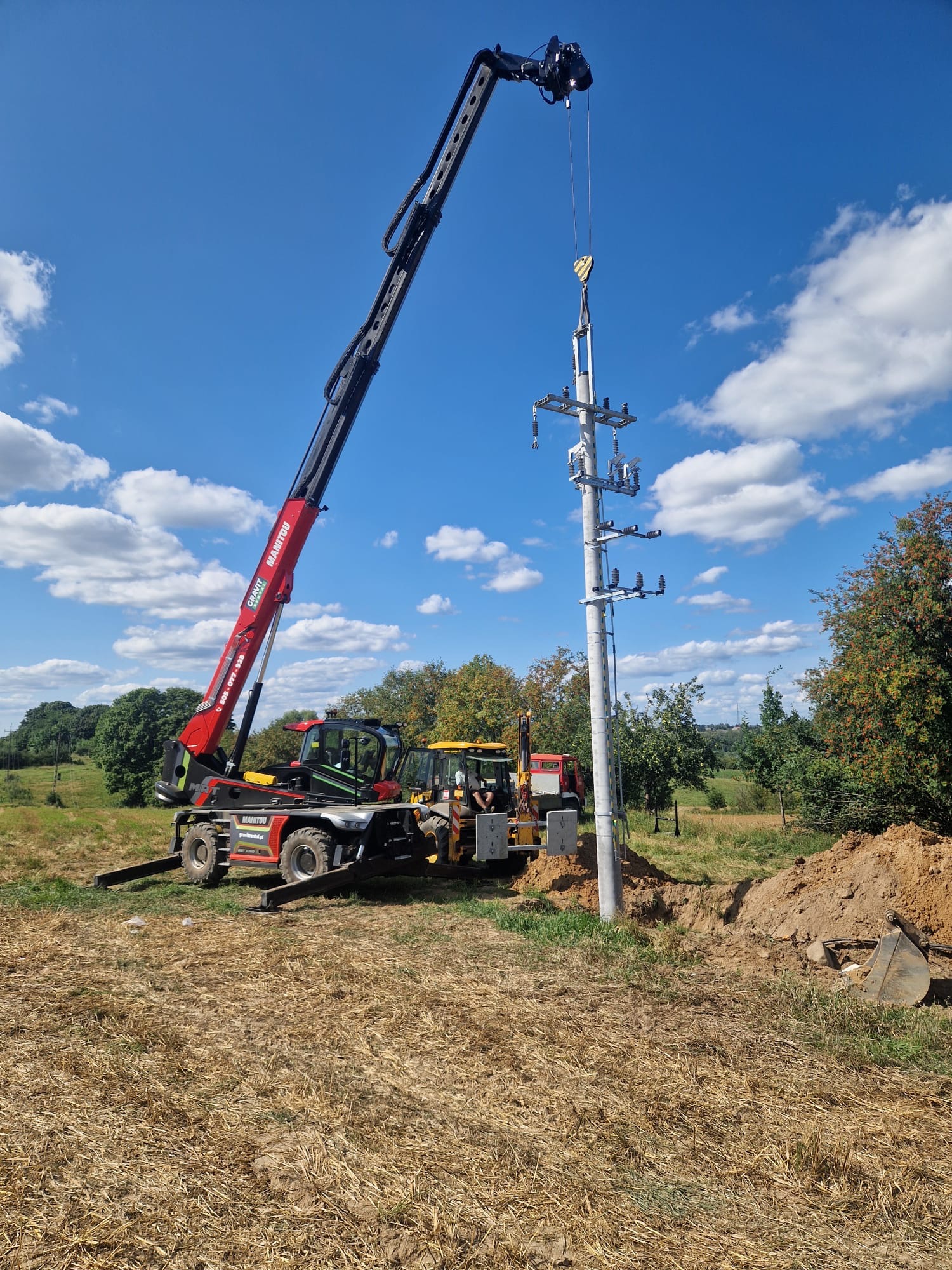 Montaż słupa energetycznego z osprzętem przy użyciu dźwigu Manitou na tle błękitnego nieba z chmurami.