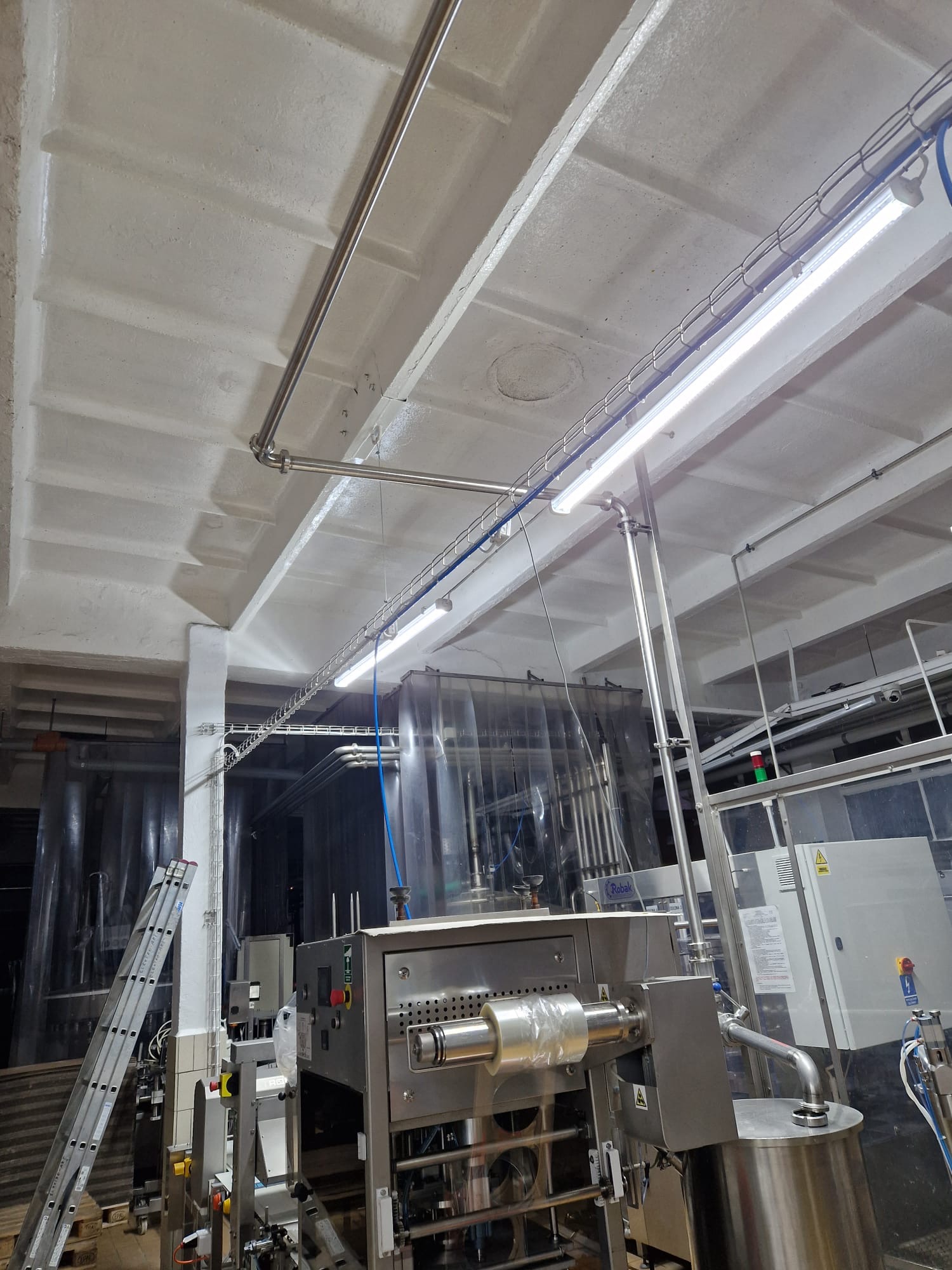 Wnętrze hali produkcyjnej z zainstalowanym oświetleniem LED, widoczne rury ze stali nierdzewnej, maszyny przemysłowe oraz drabina oparta o ścianę.