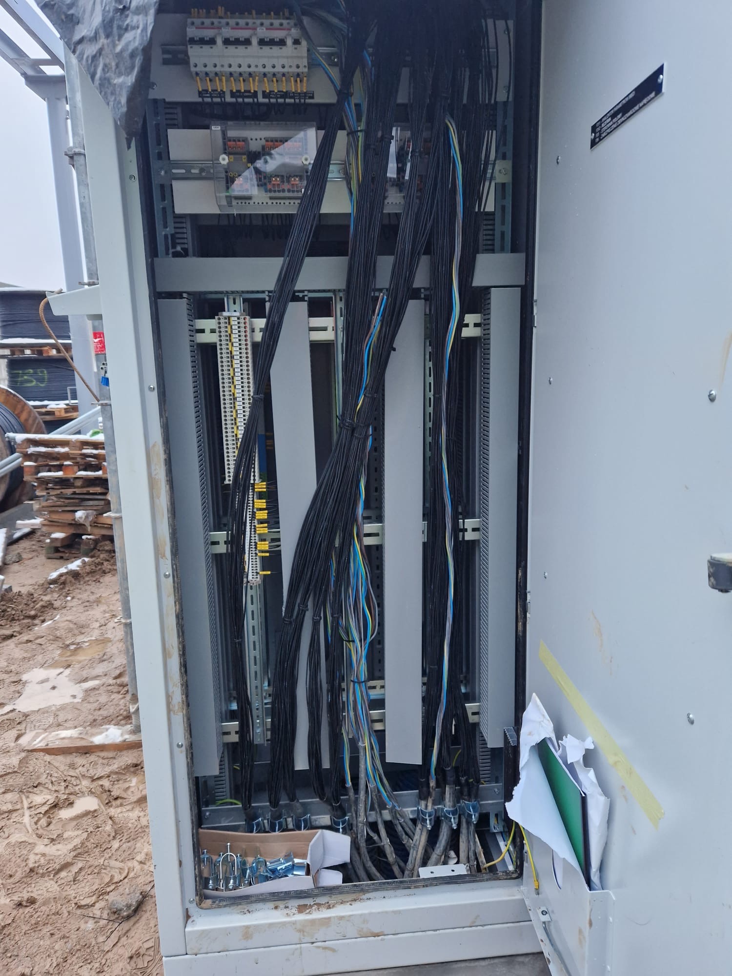 Otwarta szafa sterownicza z widocznymi kablami, listwami zaciskowymi i elementami montażowymi, na zewnątrz błotniste podłoże.