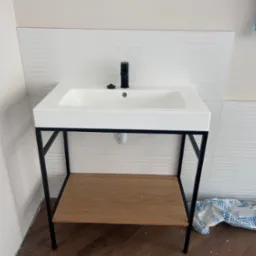 Nowoczesna, biała umywalka z czarną armaturą i metalowym stelażem z drewnianą półką, zamontowana na tle białych, poziomych płytek, z widocznymi śladami prac instalacyjnych na podłodze.