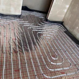 Białe rury ogrzewania podłogowego ułożone na folii z czerwoną kratką, przymocowane czarnymi klipsami, widoczne przygotowania do zalania wylewką.