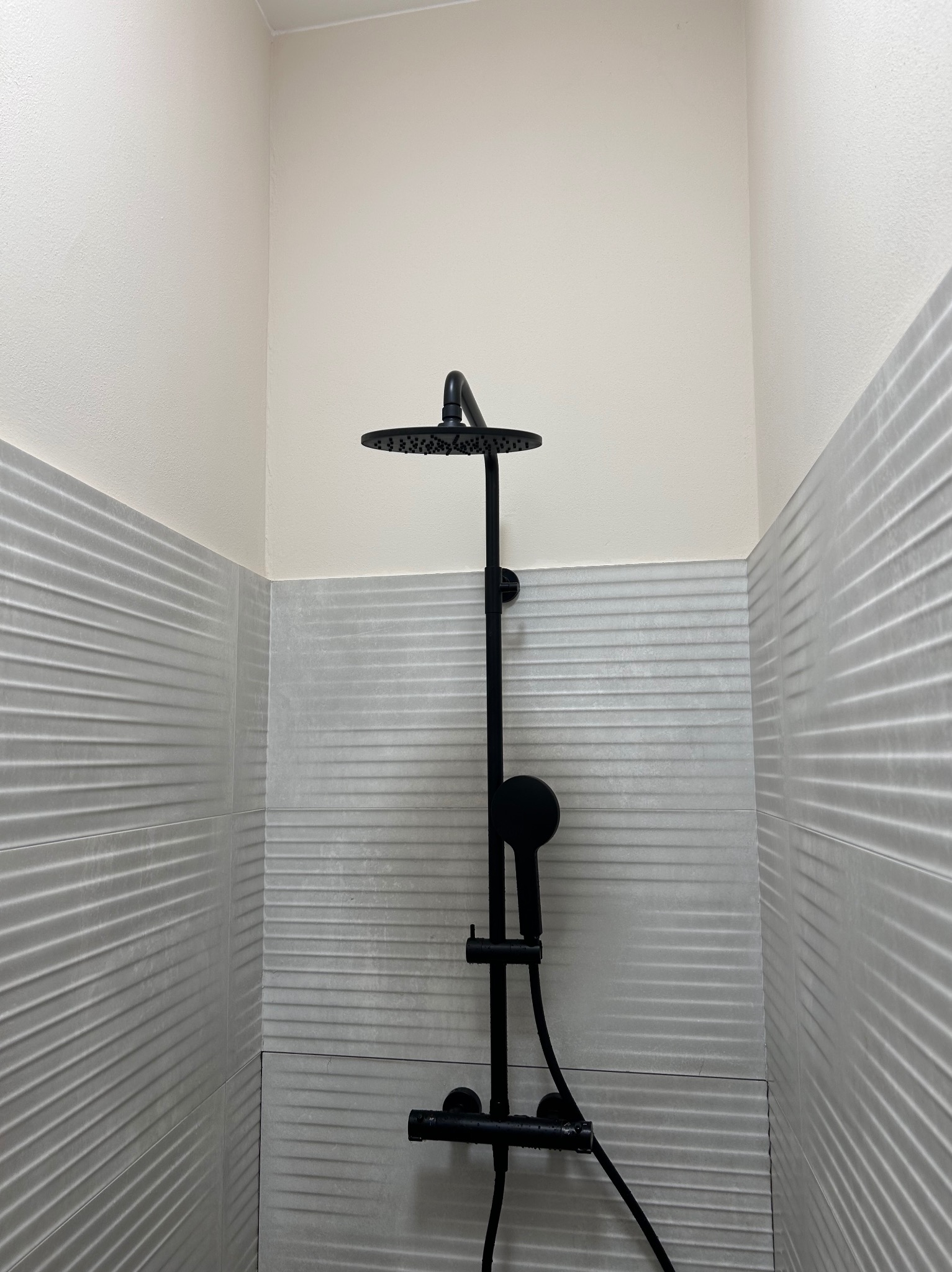 Czarny zestaw natryskowy z deszczownicą zamontowany na ścianie z szarych płytek o falistej strukturze w kabinie prysznicowej.