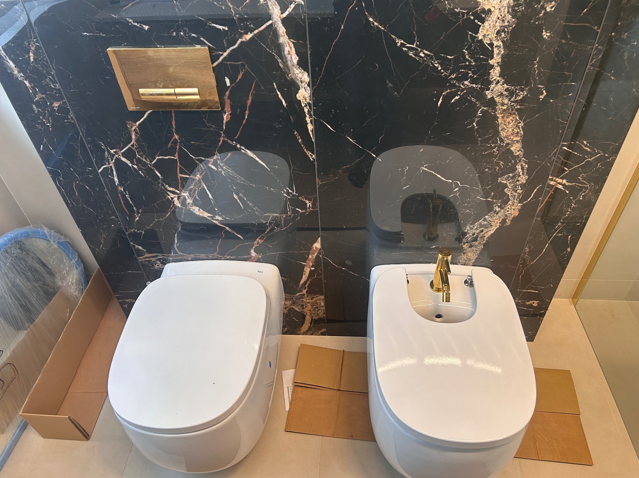 Biała toaleta i bidet zamontowane na tle ściany z czarnego marmuru z złotymi żyłami, widoczny złoty przycisk spłuczki i kartonowe zabezpieczenia pod urządzeniami.