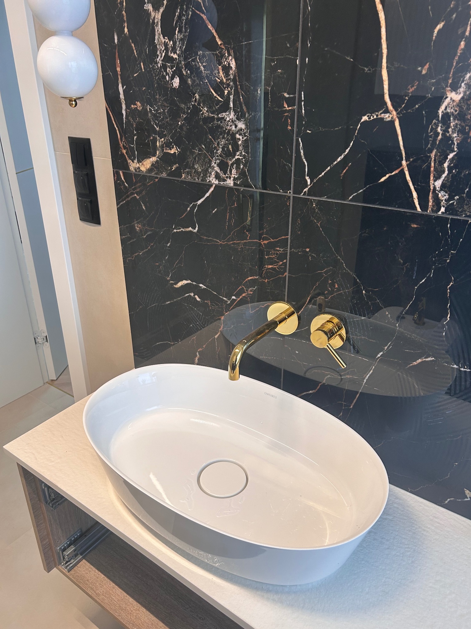 Nowoczesna łazienka: biała, owalna umywalka nablatowa z złotą armaturą na tle ściany z czarnego marmuru z złotymi żyłami. Widoczne elementy szafki pod umywalką oraz włączniki światła.
