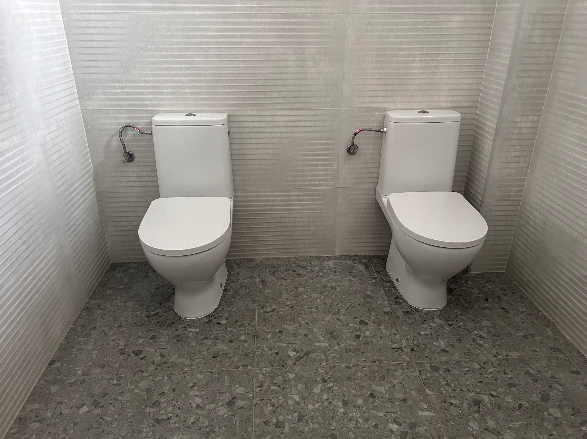 Dwa białe kompakty WC zamontowane obok siebie na podłodze z szarej terazzo, z widocznymi przyłączami wody i białymi płytkami ściennymi z poziomymi żłobieniami.
