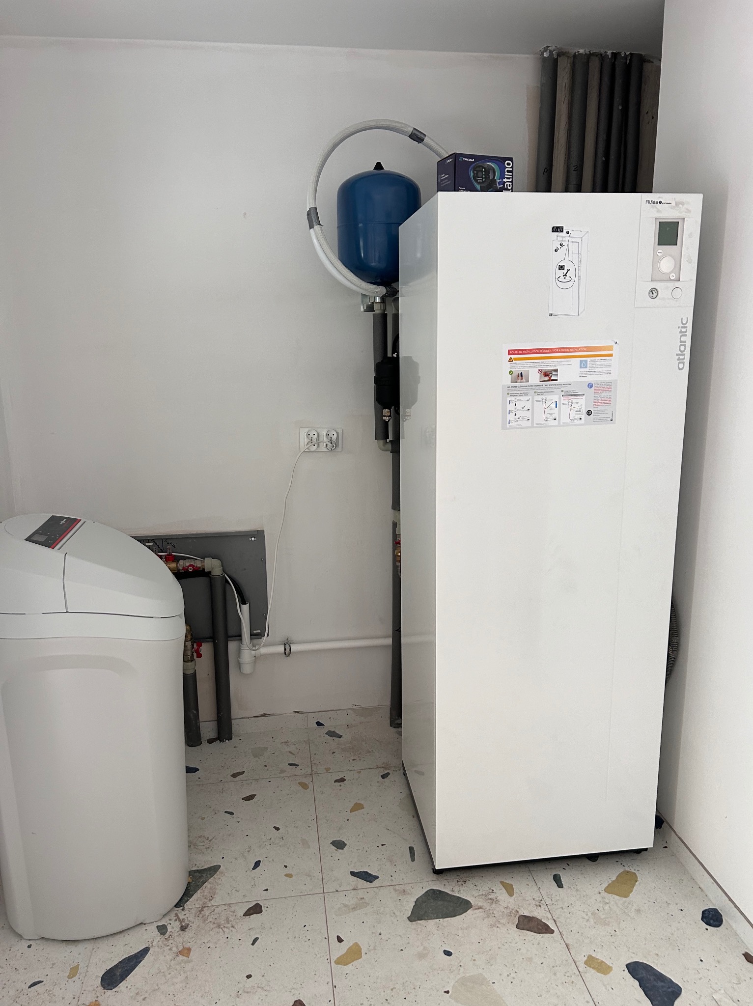 Biała pompa ciepła Atlantic z niebieskim zbiornikiem wyrównawczym i zmiękczaczem wody w pomieszczeniu z podłogą w stylu lastryko.