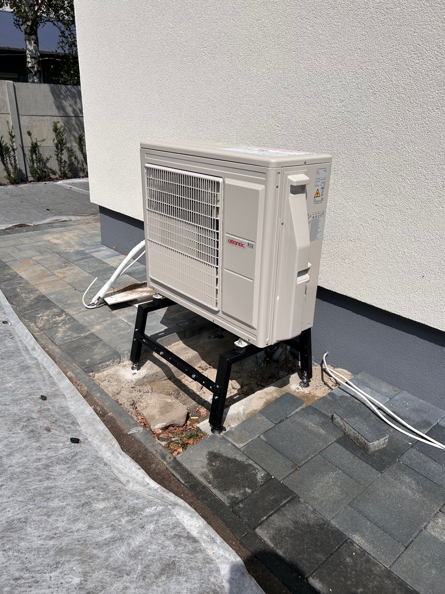 Zewnętrzna jednostka klimatyzacji marki Atlantic zamontowana na czarnym metalowym stelażu przy ścianie budynku, widoczne przewody instalacyjne na kostce brukowej.