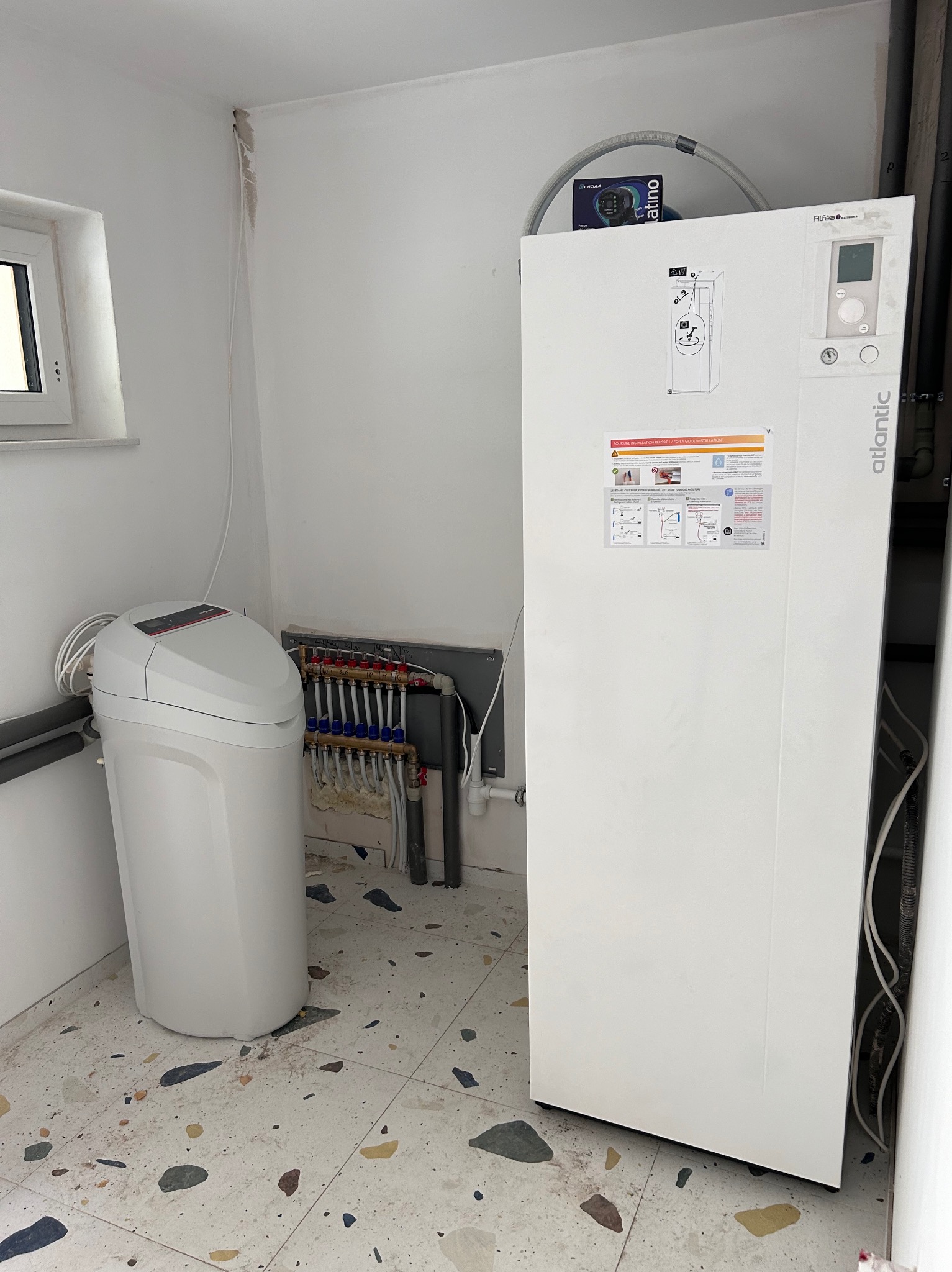 Biała pompa ciepła Atlantic oraz zmiękczacz wody w pomieszczeniu z terakotą, widoczne rury instalacji grzewczej i fragment okna.