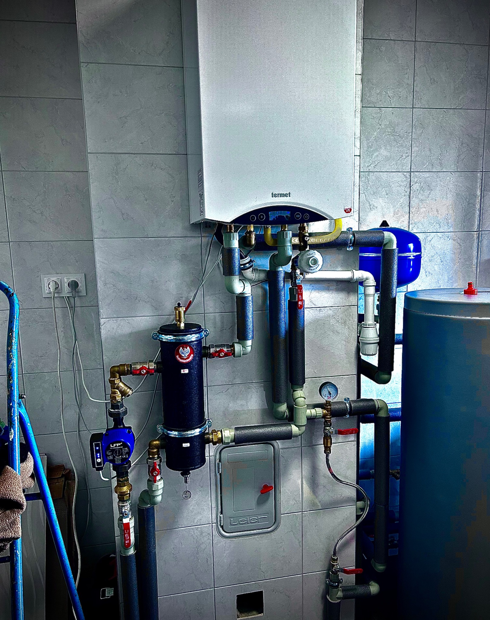 Kocioł gazowy marki Termet z podłączonymi rurami, zbiornikiem wyrównawczym i pompą obiegową, zamontowany na ścianie wyłożonej szarymi płytkami.