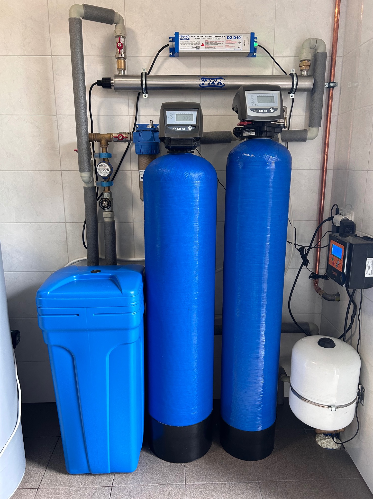 Dwa niebieskie filtry do wody z elektronicznymi wyświetlaczami, obok niebieski pojemnik na sól, instalacja z rur miedzianych i szarych, lampa UV do sterylizacji wody, zamontowane w pomieszczeniu...